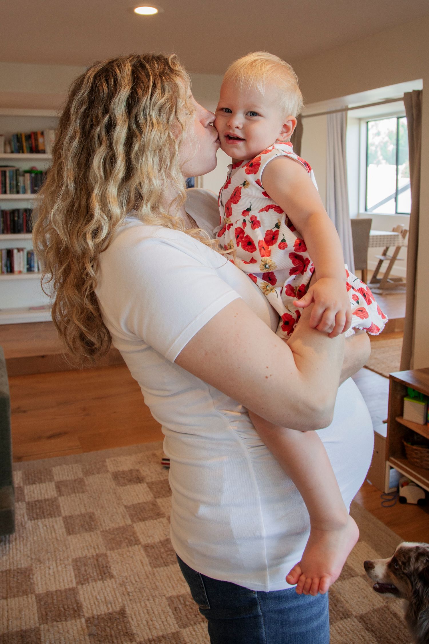 A baby in a floral dress being held and comforted in a living room with bookshelves and carpeting.