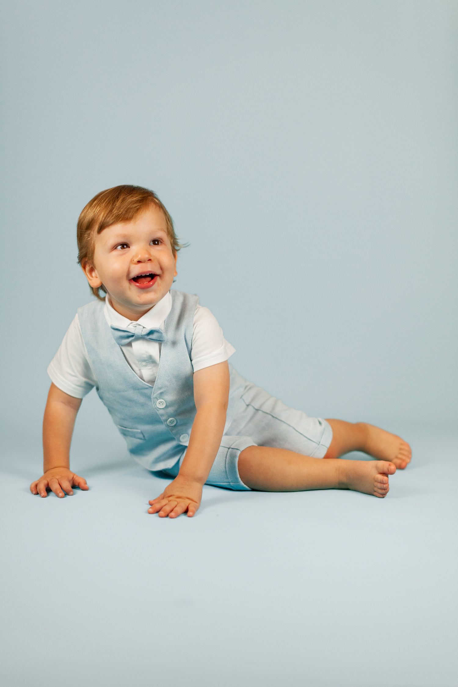 Baby in light blue outfit lying on stomach and laughing joyfully against pale blue studio background.