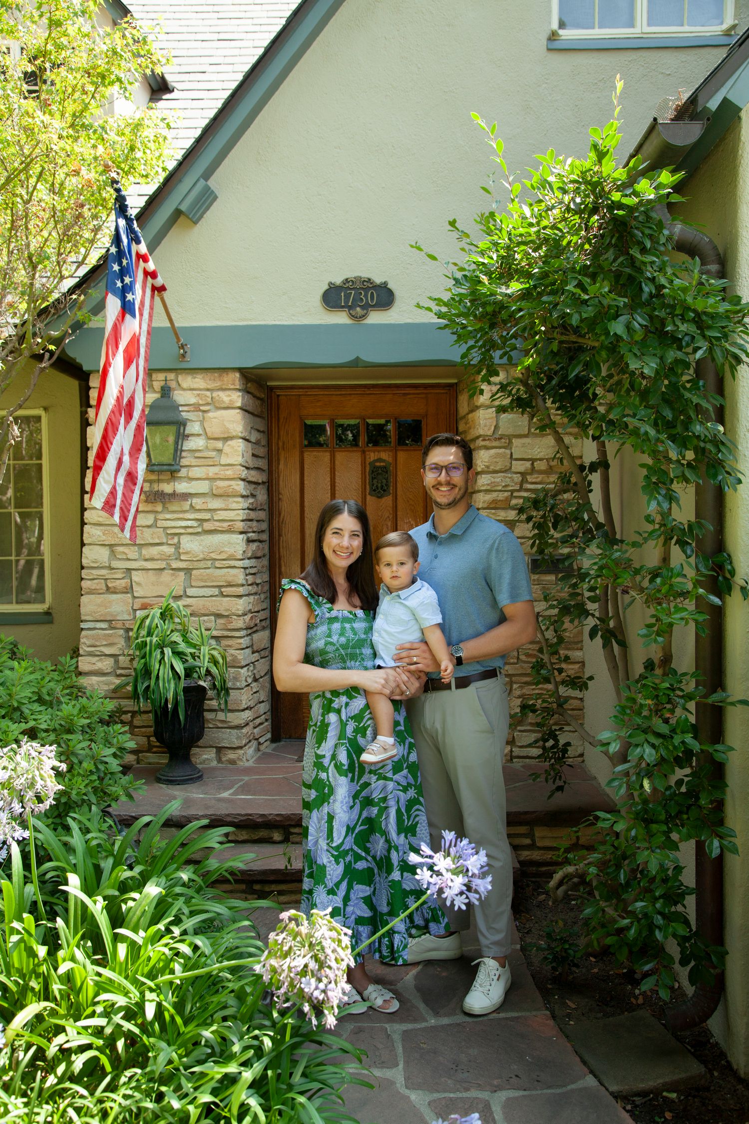 A family poses together on a stone porch with American flags and greenery decorating the entrance of their home.