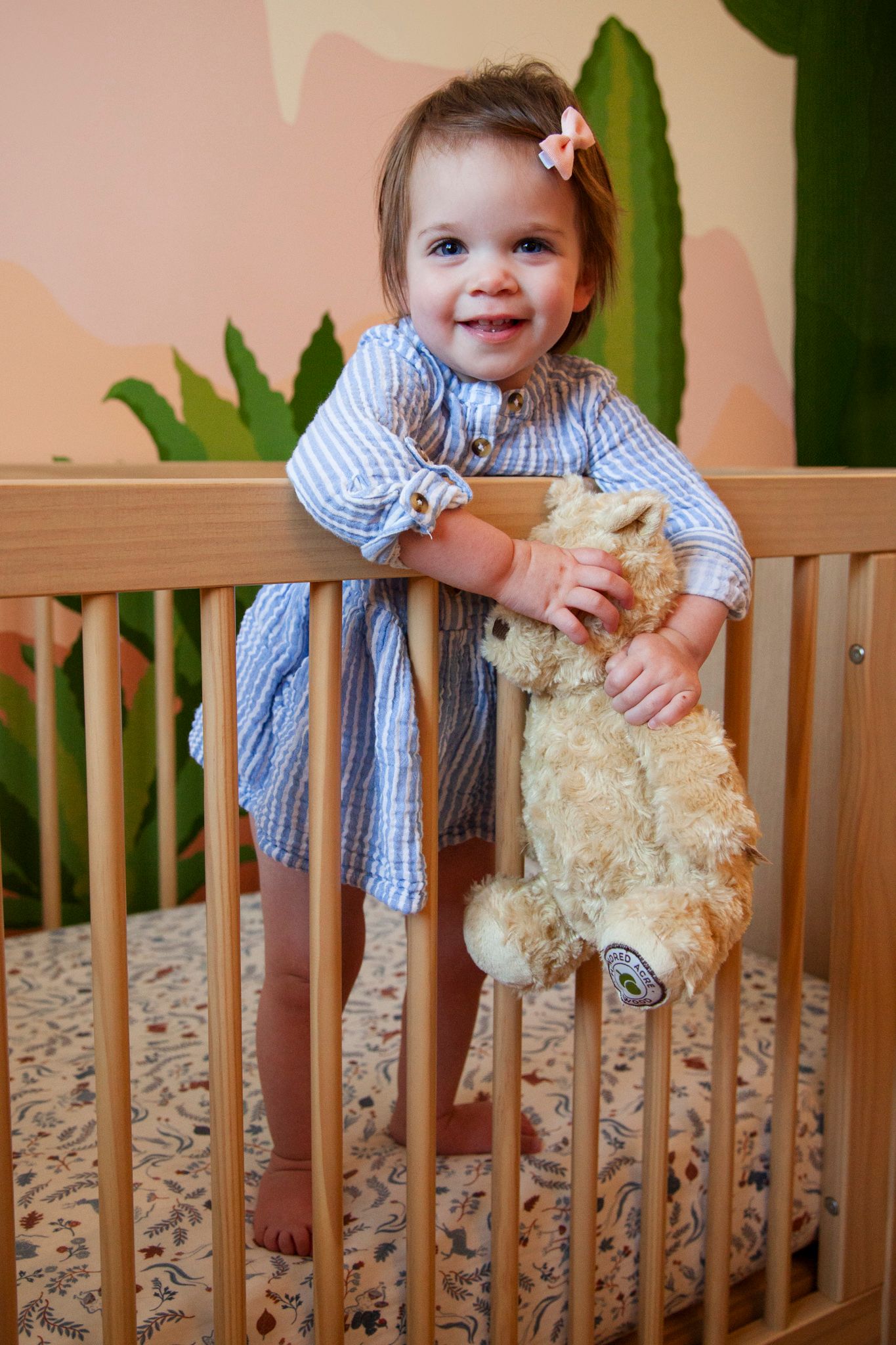Baby in blue gingham shirt stands in wooden crib holding stuffed animal toy in nursery setting.