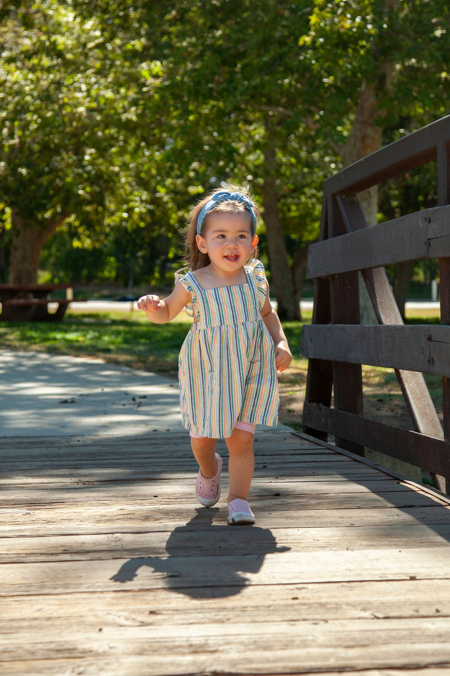 A toddler in striped outfit walks happily along a wooden bridge pathway in a sunny park setting.