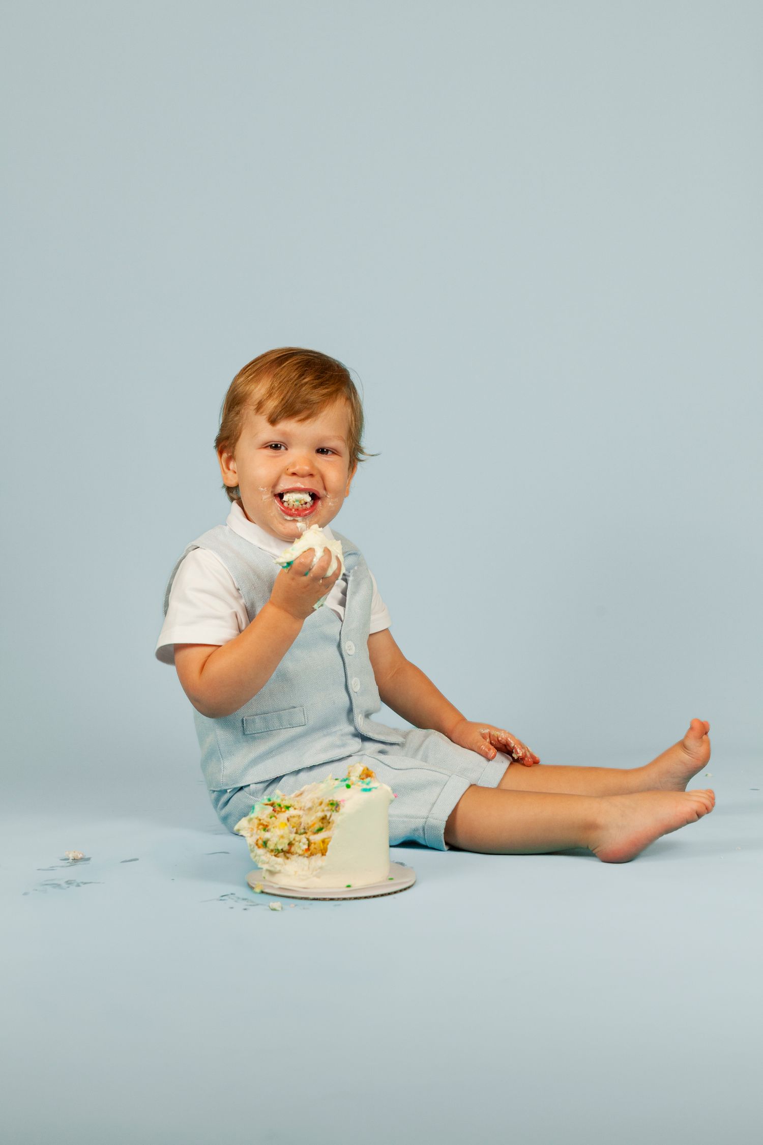 A small child in white clothing enjoying cake and treats against a light blue background.