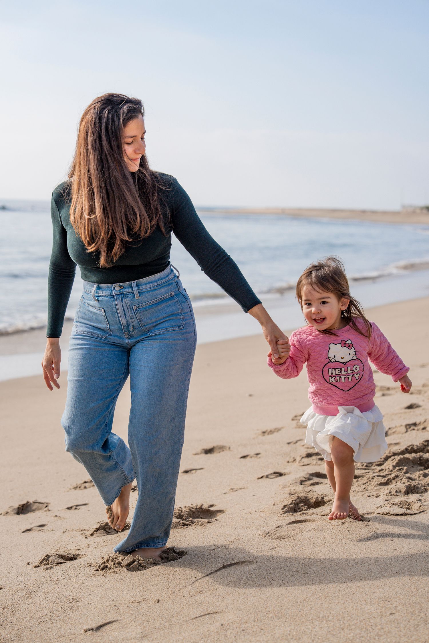 A parent and child walk along a sandy beach shoreline in casual clothing on a sunny day.