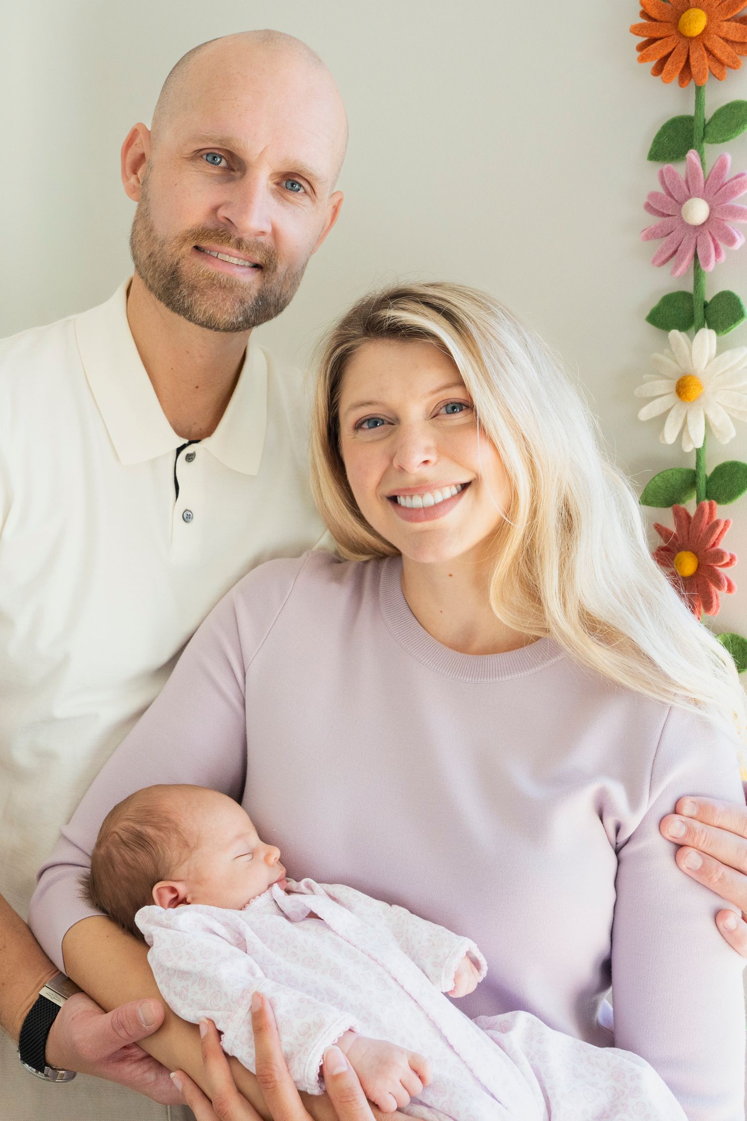 A couple in white and lavender poses against a floral backdrop with their newborn baby in a series of tender moments.
