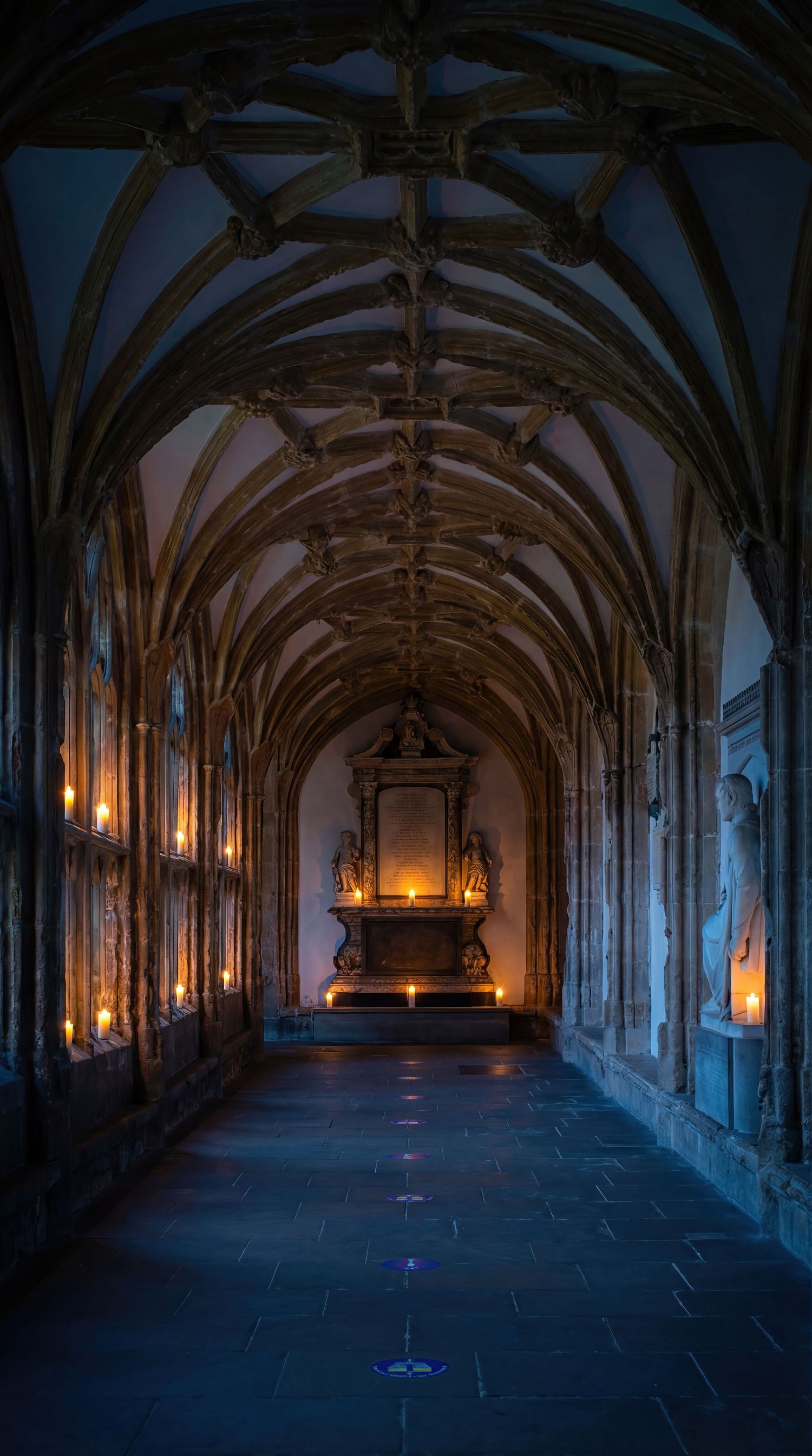 Gothic stone corridor with vaulted ceiling and glowing wall sconces creates dramatic lighting in a historic monastery.