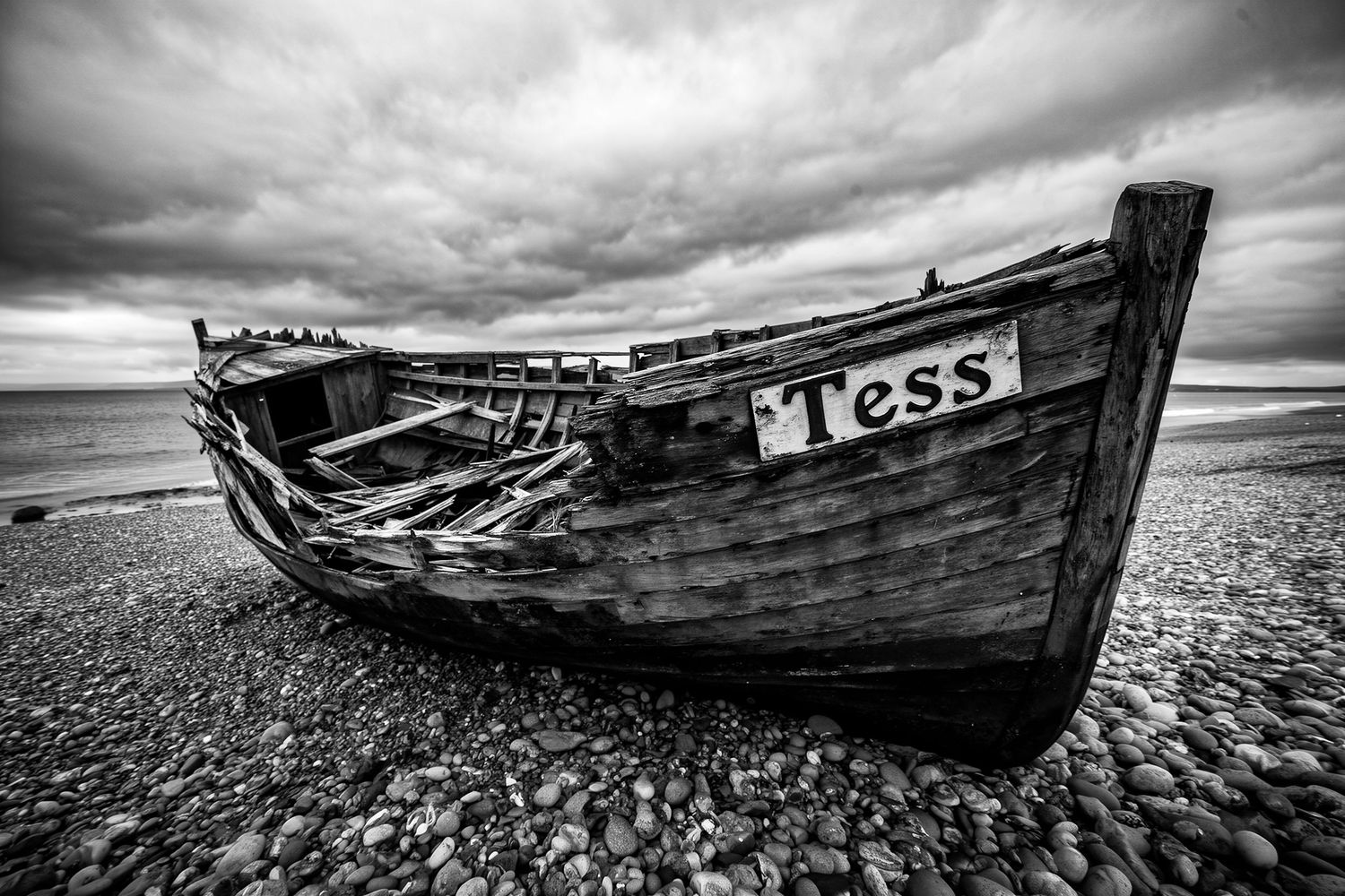 Black and white photo of an abandoned wooden boat named Tess resting on a rocky beach under dramatic cloudy skies.