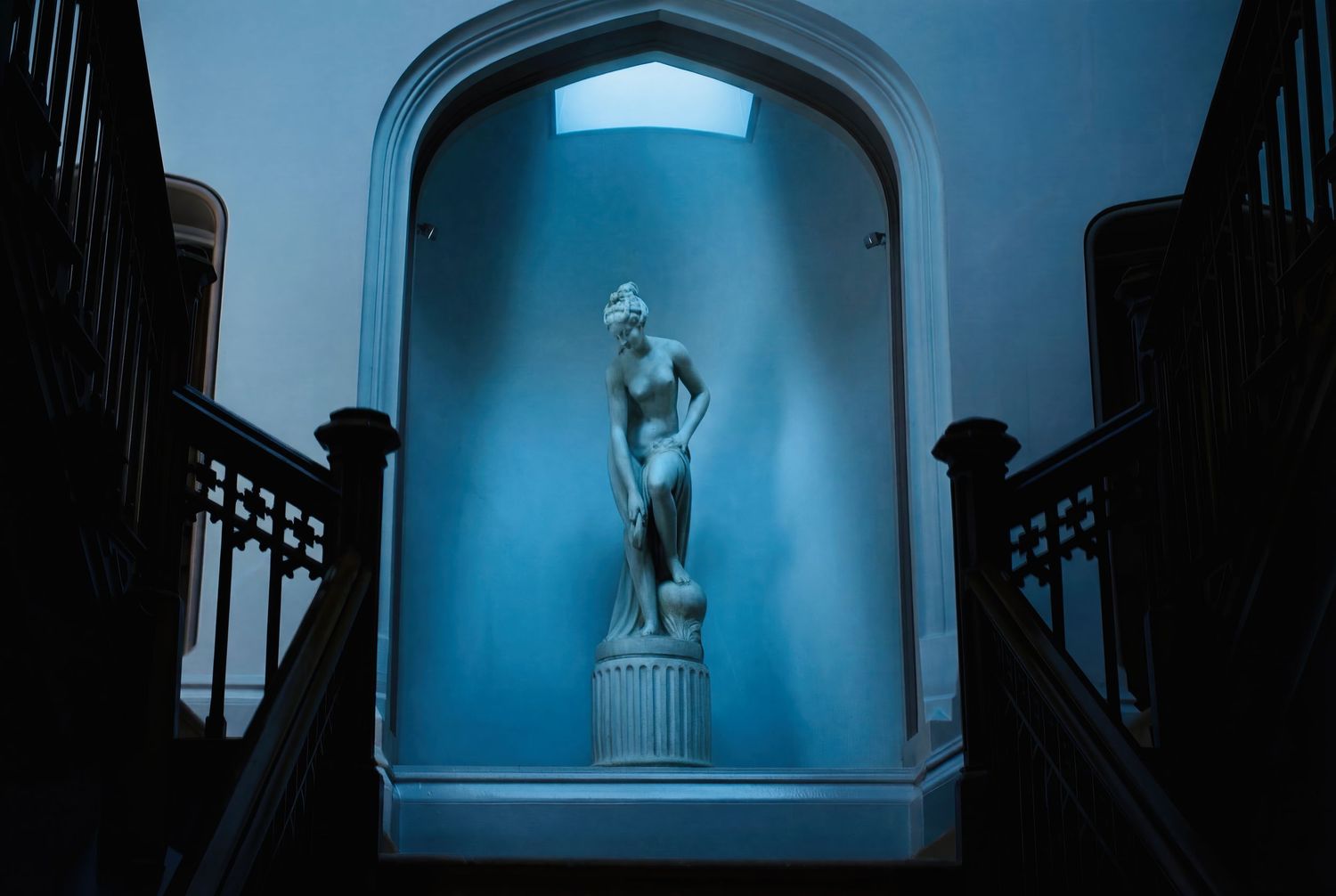 Dramatic blue-lit arched hallway with classical statue silhouetted at the end creates moody architectural atmosphere.