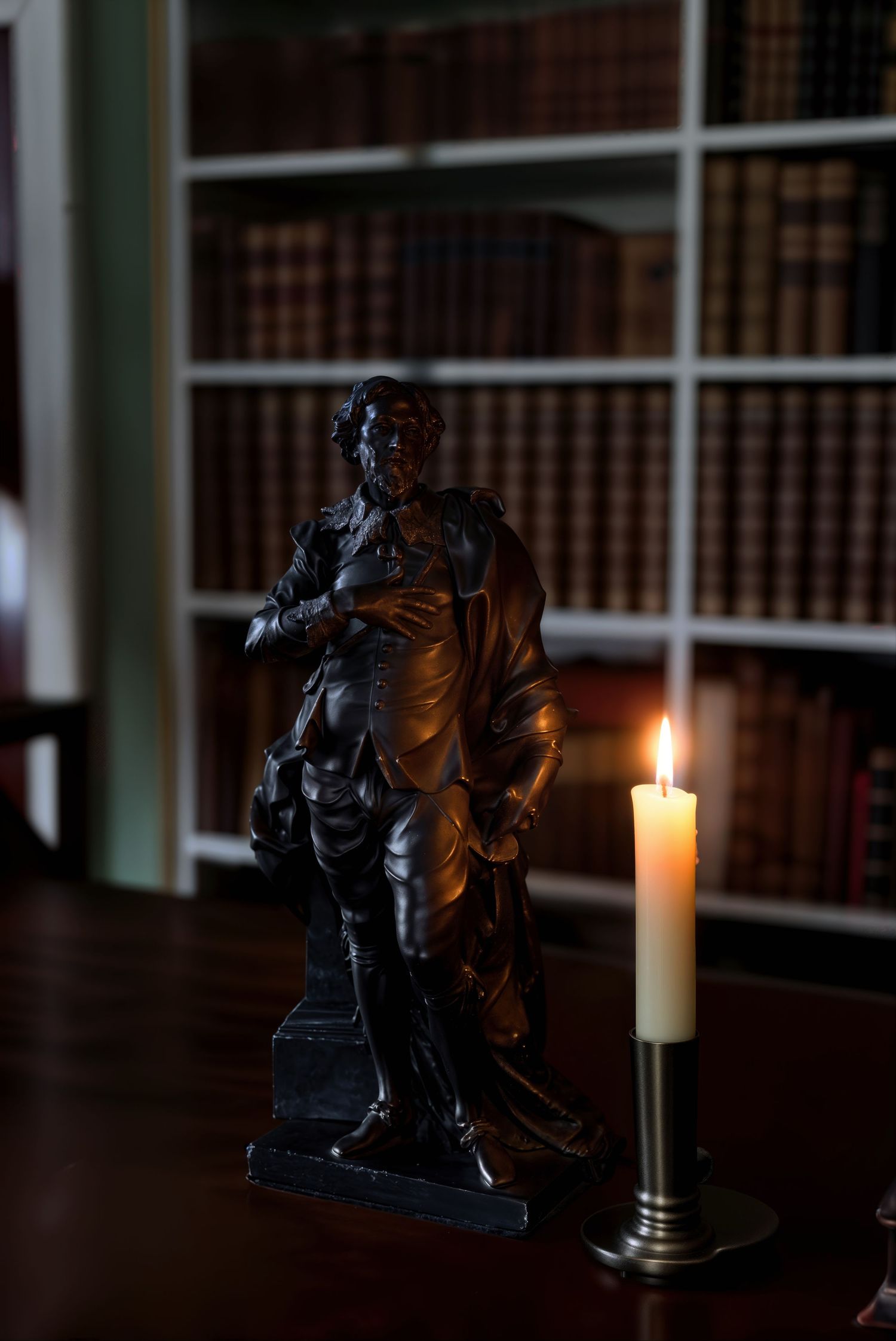 Dark bronze statue of a historical figure beside a lit candle on a wooden desk in a classic library.