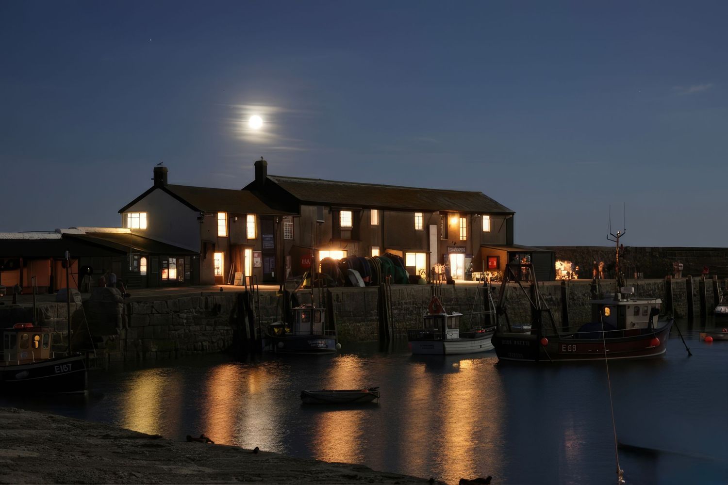 Moonlit harbor at night with glowing fishing boats reflected in calm water beside a stone building.