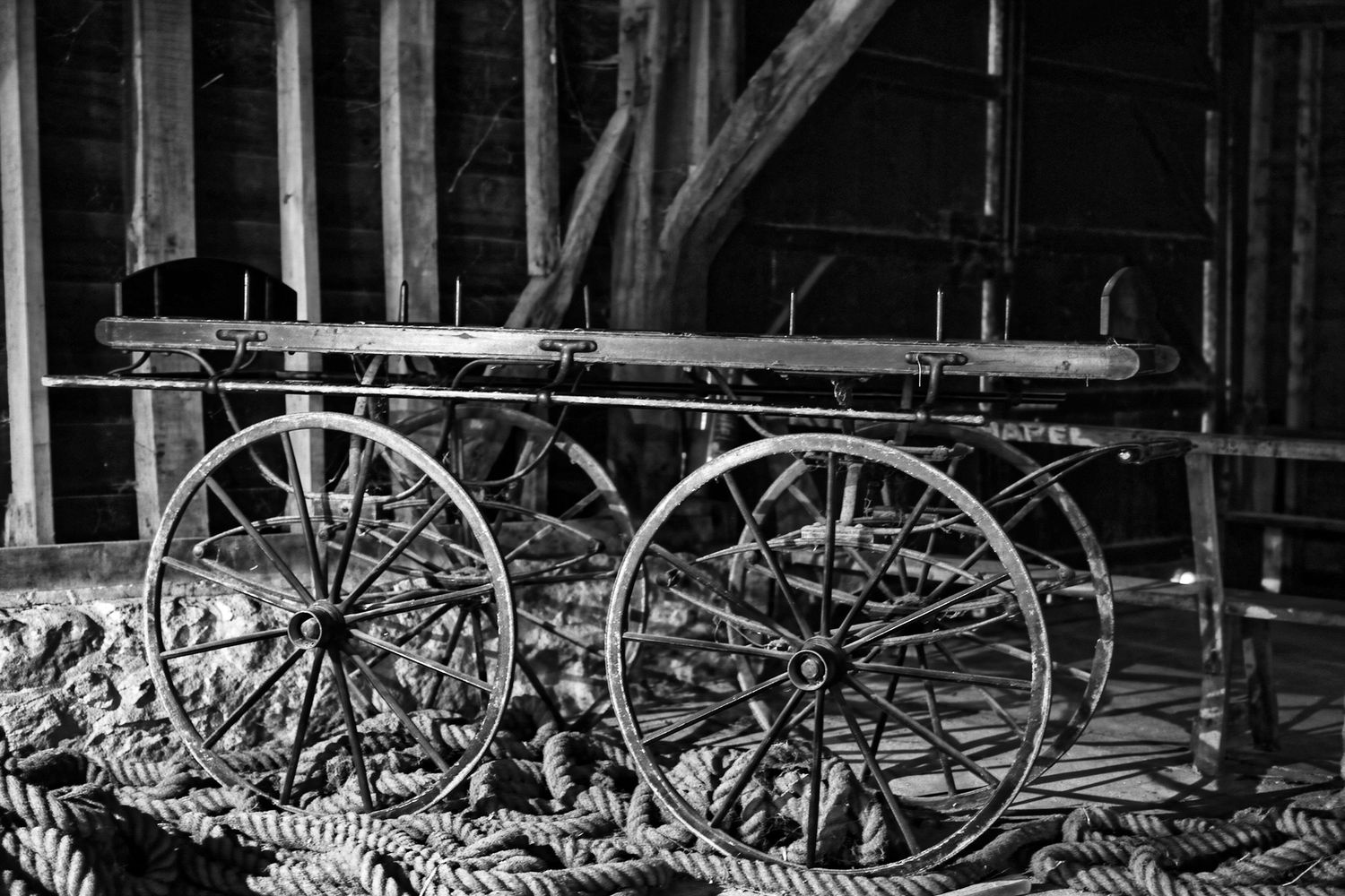 Vintage black and white photo of antique wagon wheels and wooden cart inside a rustic barn with rope.