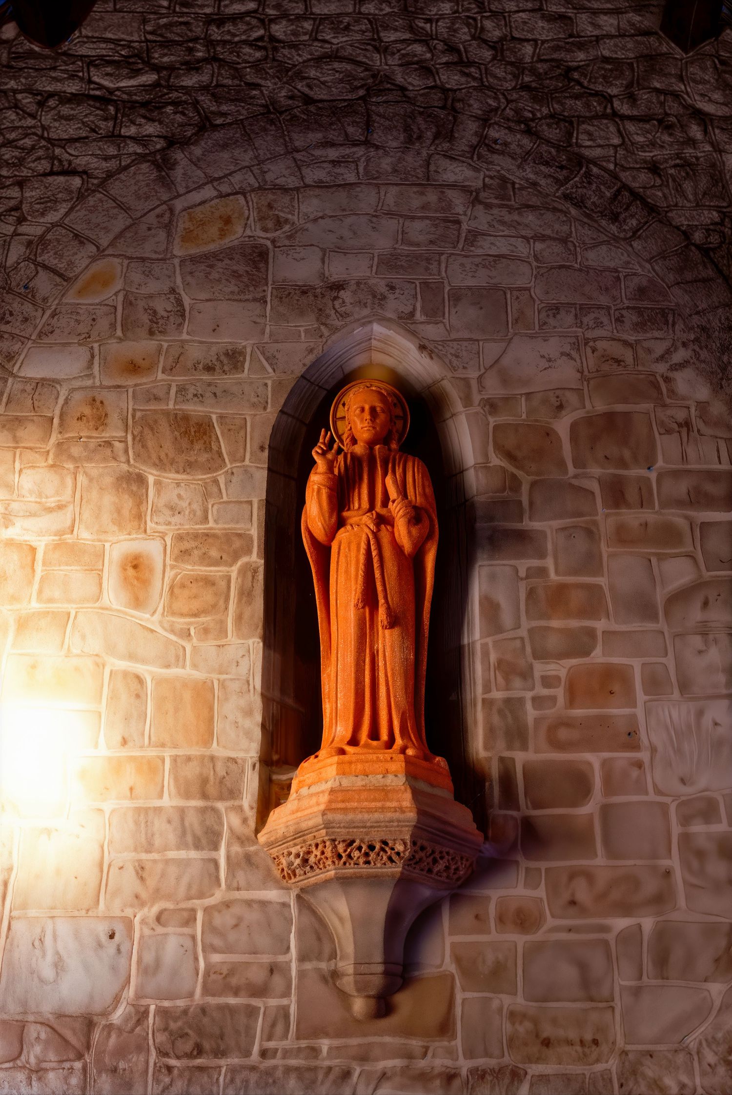 Stone niche in a church wall featuring an illuminated religious statue bathed in warm orange light.