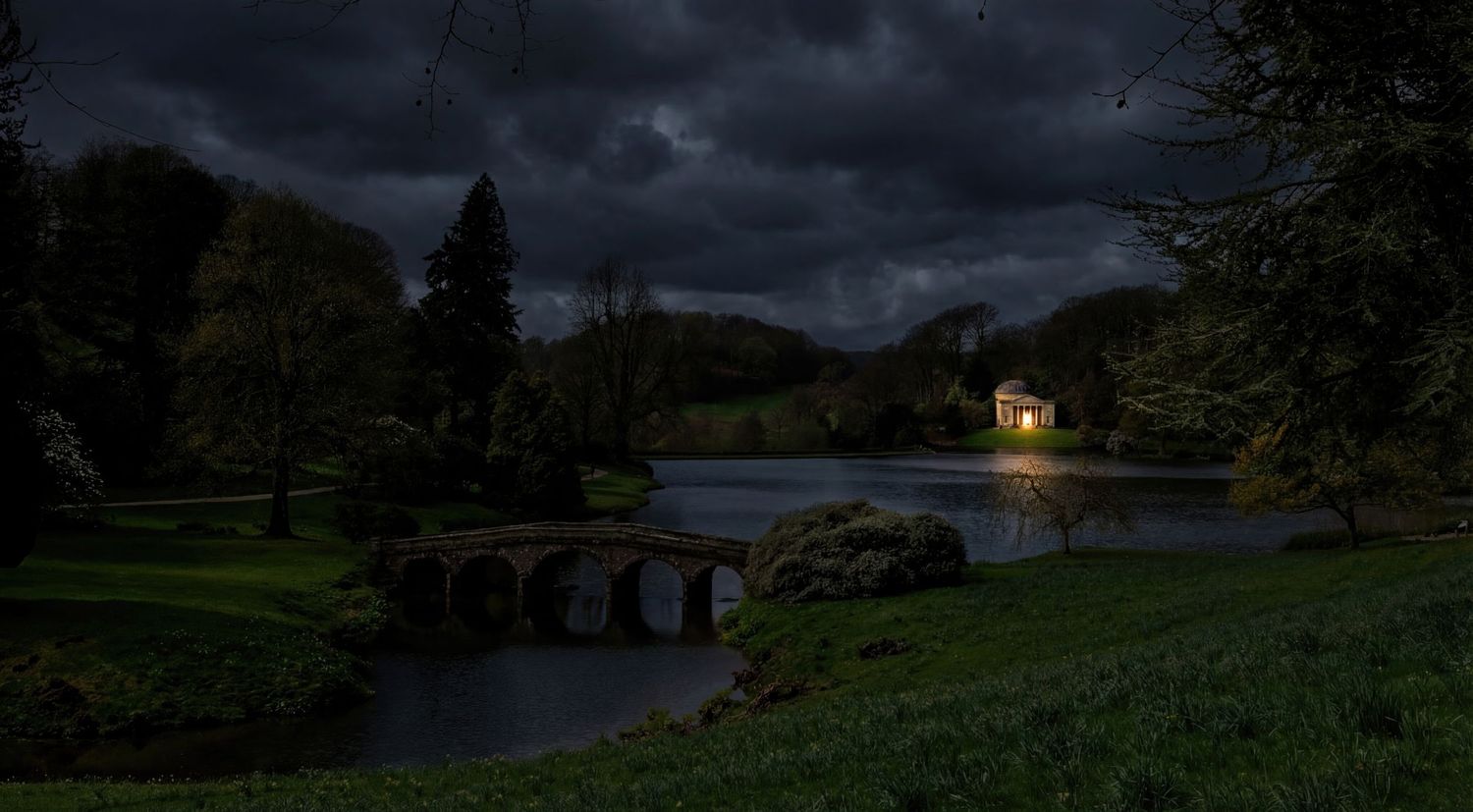 Dark moody landscape of a lakeside path at dusk with distant lights glowing through trees.