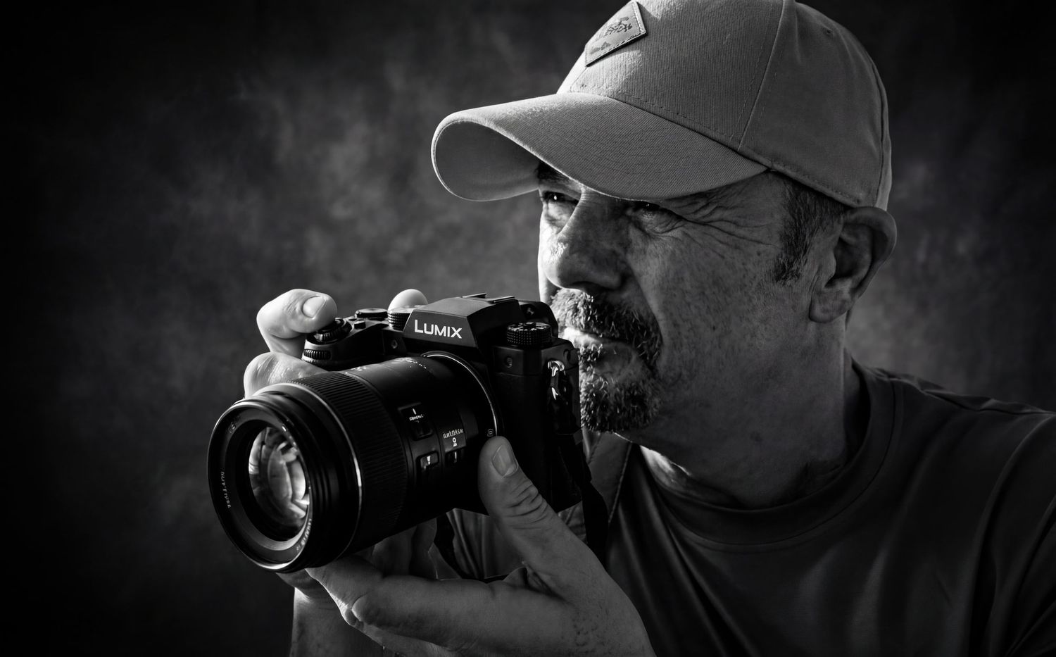 Black and white portrait of a man wearing a cap, holding a Lumix camera up to his eye while photographing.