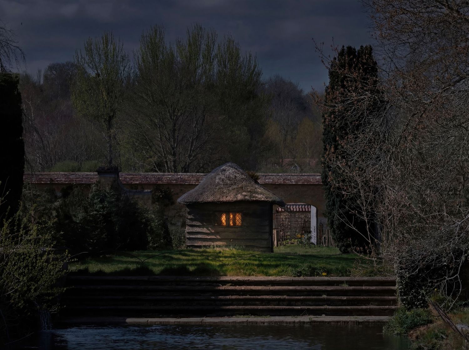 A rustic log cabin with glowing windows sits nestled among trees at dusk, with stone steps leading to the entrance.