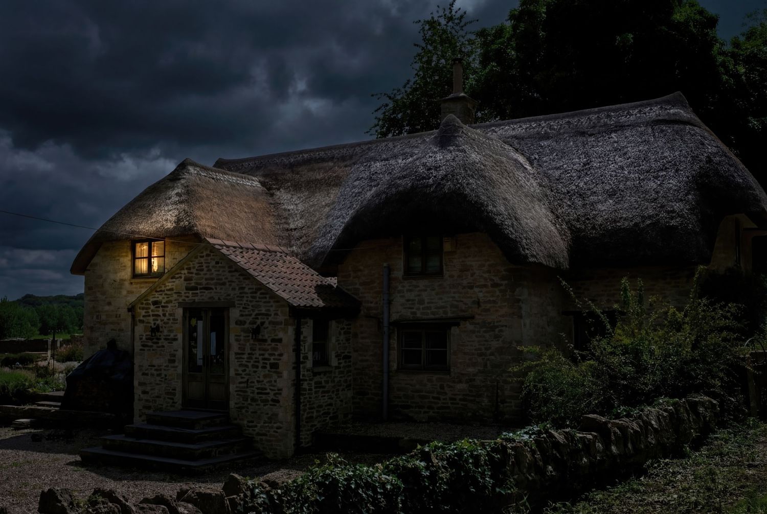 A cozy thatched-roof cottage glows warmly in the darkness with light from its windows against a stormy night sky.
