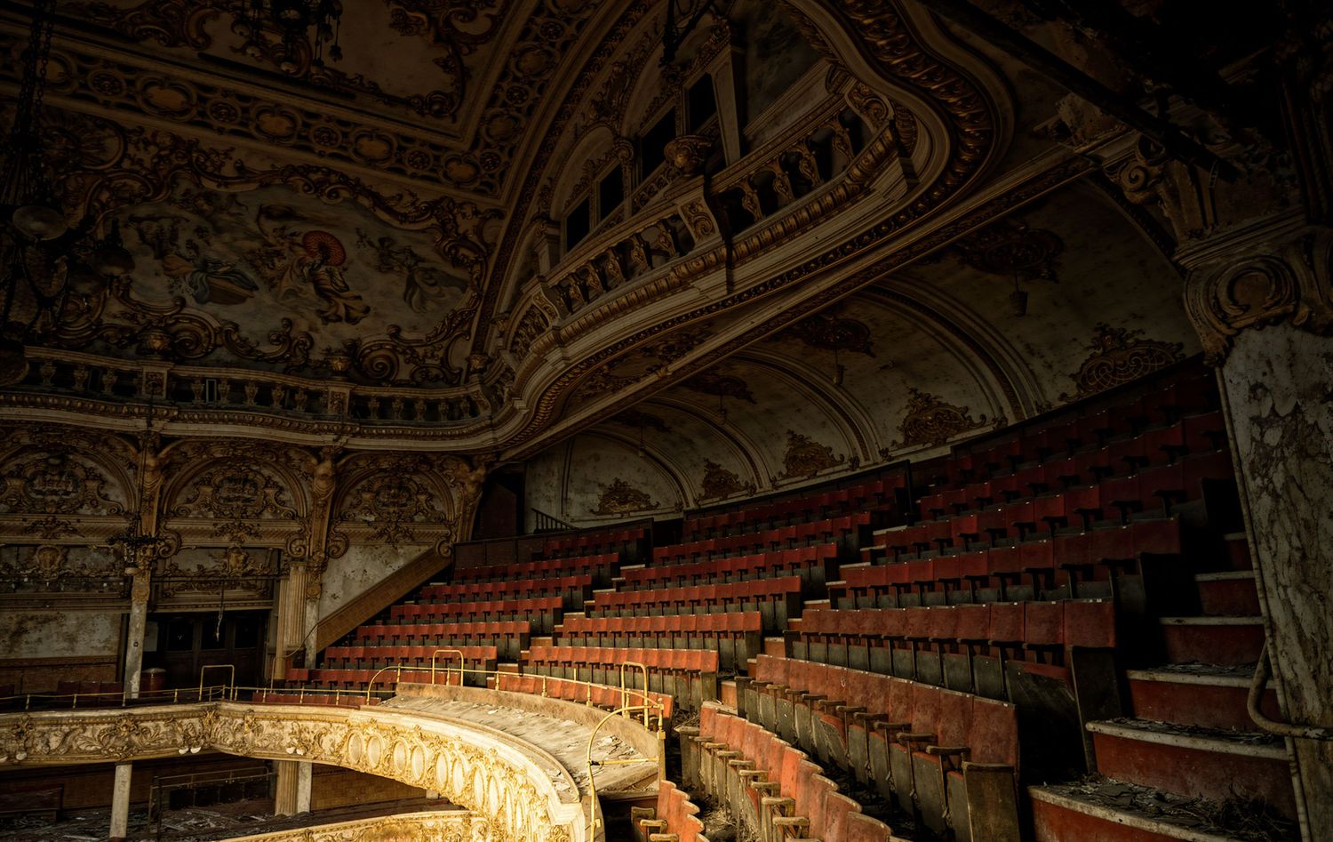 Abandoned ornate theater with rows of red seats, intricate ceiling murals, and grand balcony in decay.