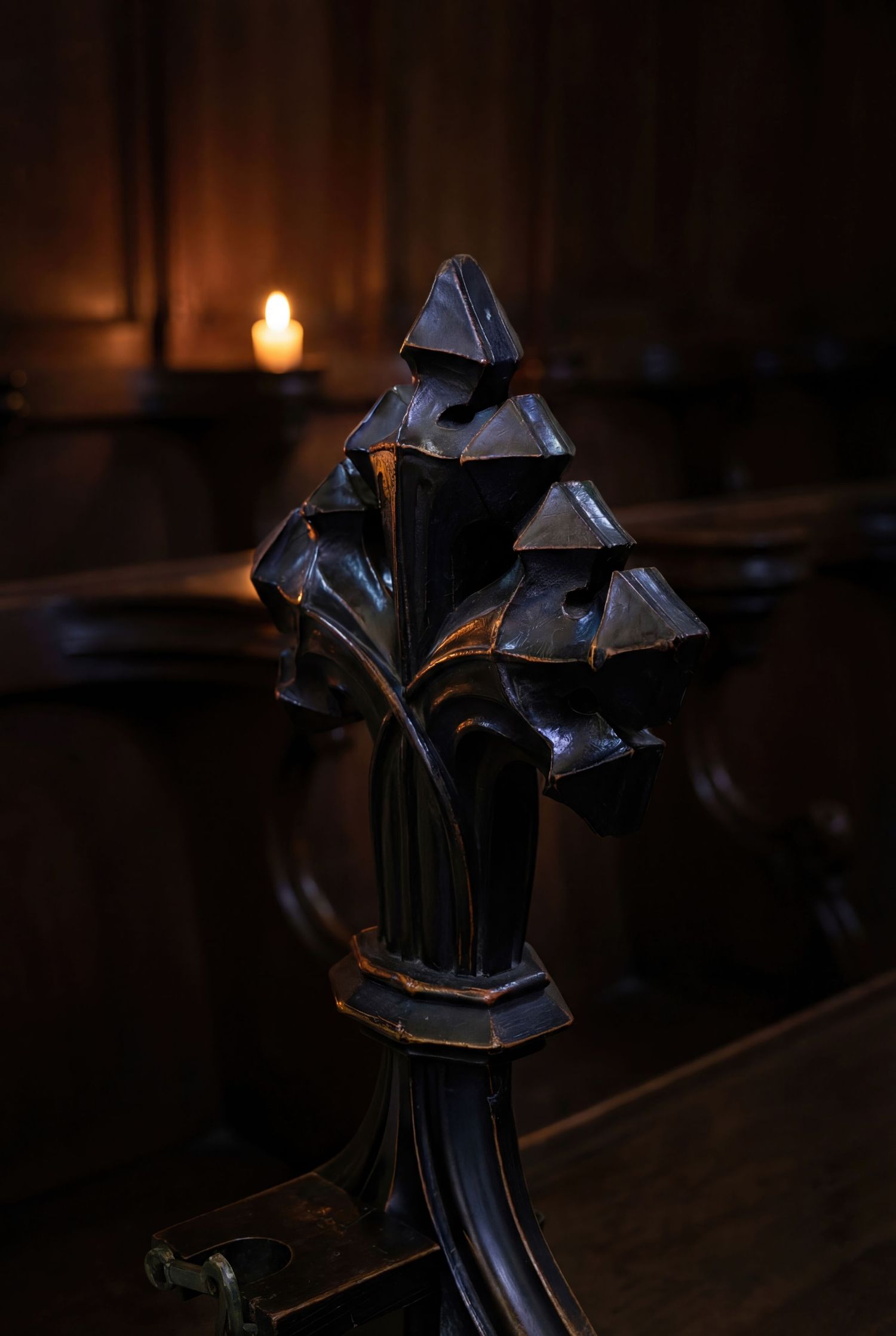 A dark ornate metal church pew end detail with a decorative finial silhouetted against a warm candlelit background.