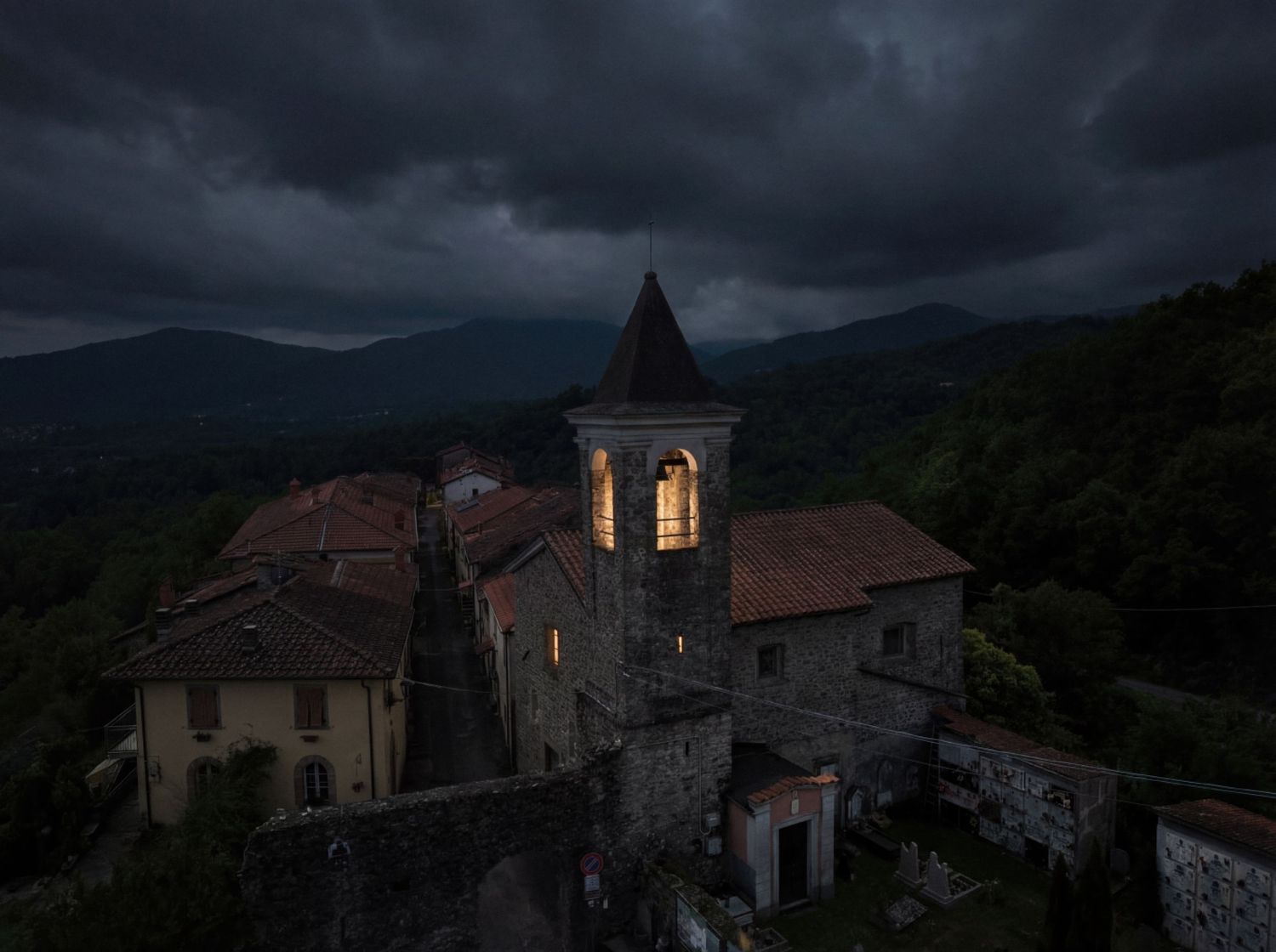Aerial view of a historic Italian village church with glowing windows under dramatic stormy skies at dusk.