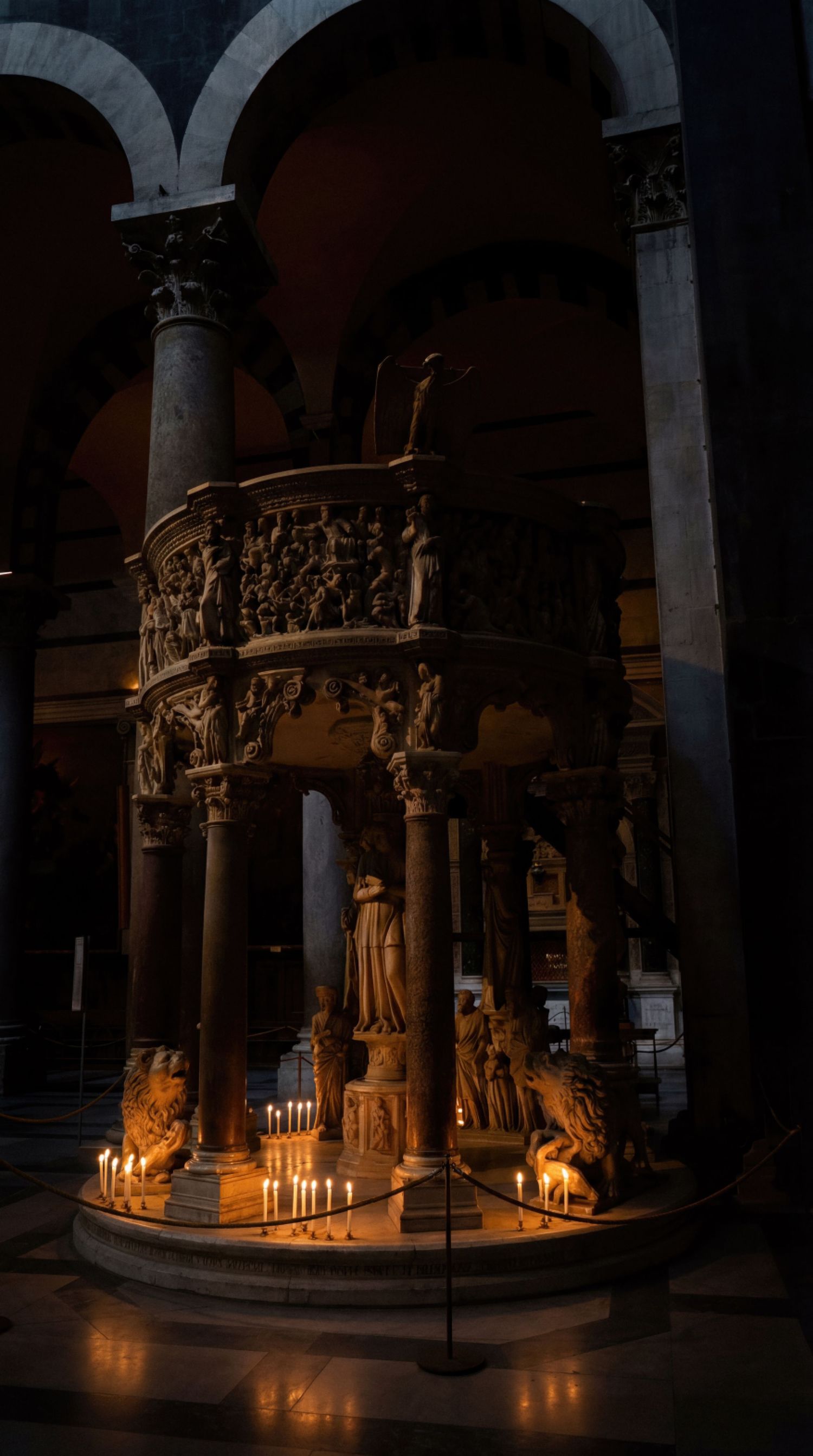 Illuminated medieval stone sculptures in a dark church cloister with arched columns and candlelight creating dramatic shadows.