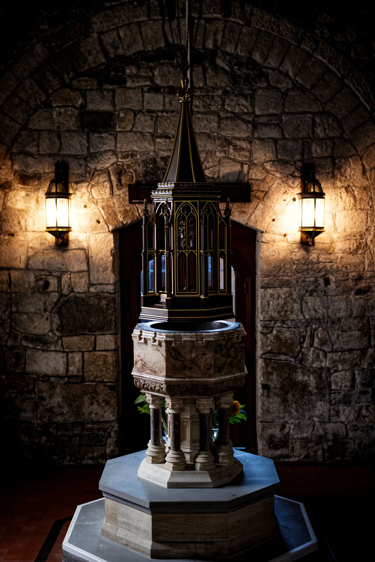 Gothic baptismal font in a medieval stone church, flanked by ornate wall lanterns beneath a arched alcove.