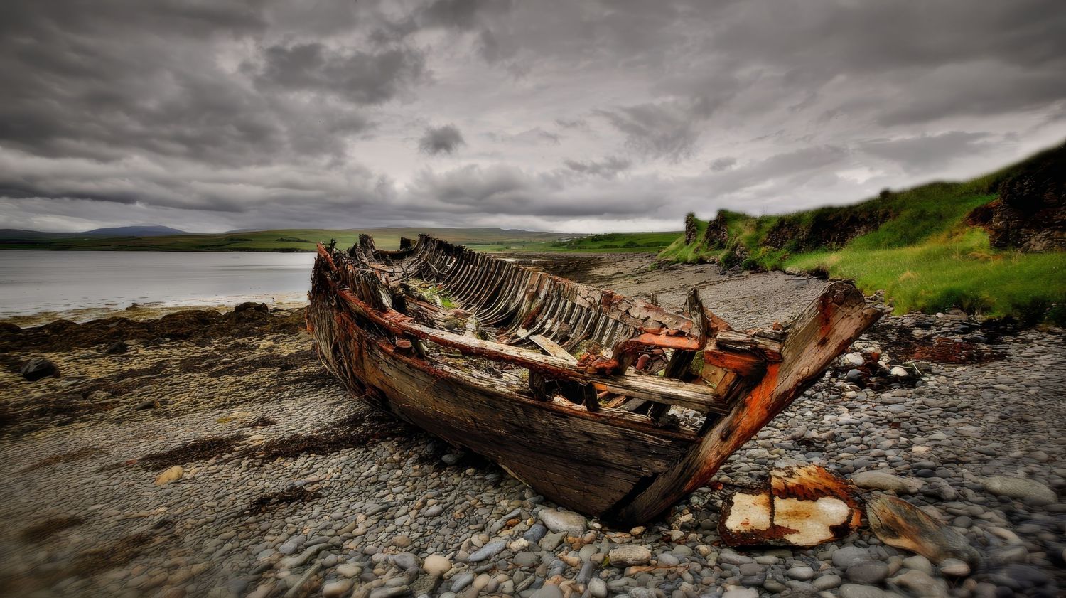 Abandoned wooden shipwreck decaying on a rocky shoreline under dramatic cloudy skies near a calm coastal inlet.
