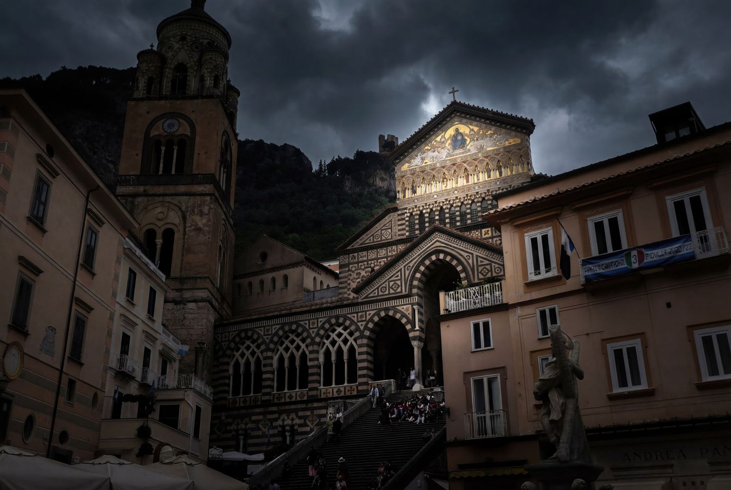 Dramatic evening view of Amalfi Cathedral in Italy with dark storm clouds and illuminated facade against historic buildings.