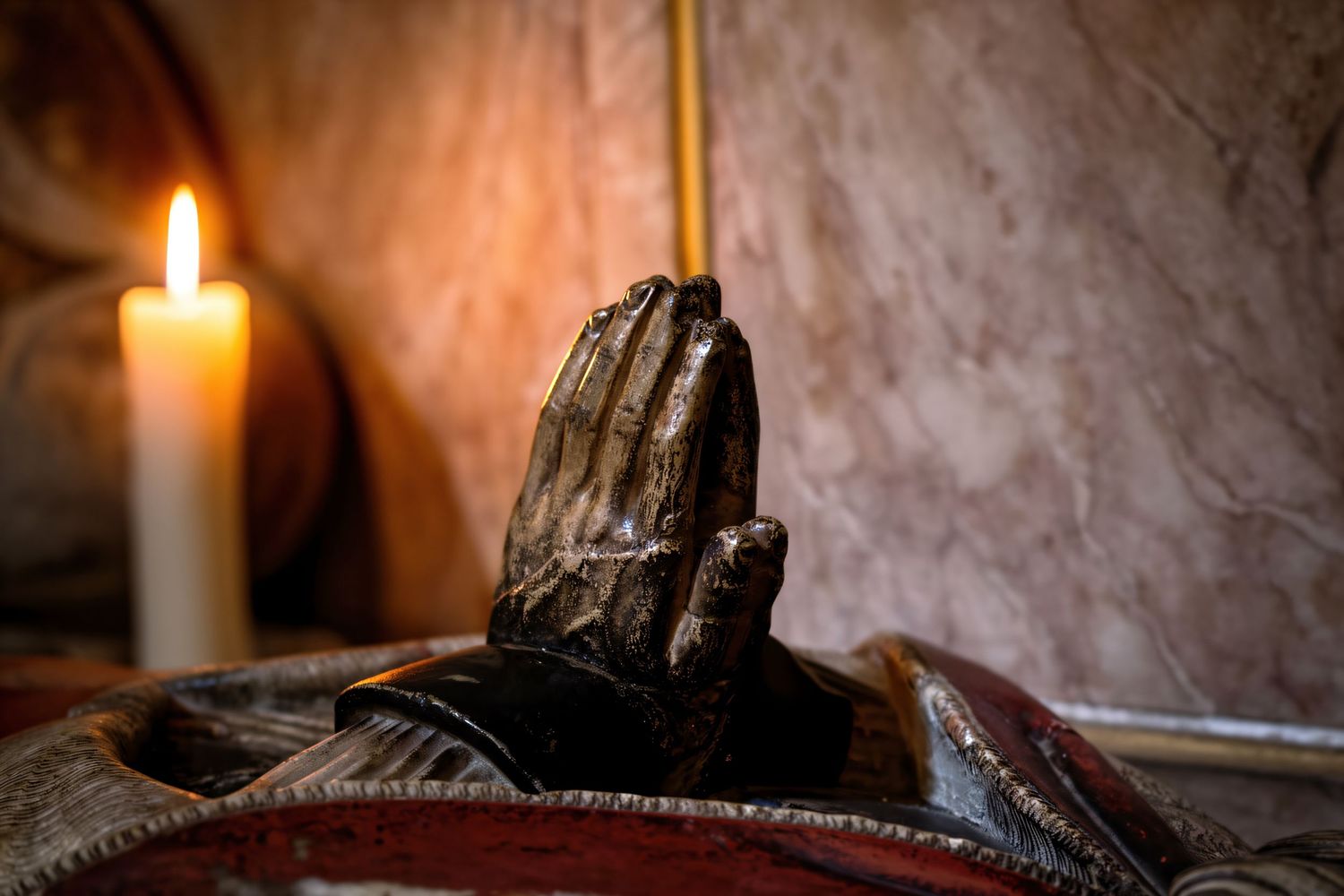 Bronze praying hands sculpture beside a lit candle in a dimly lit, atmospheric religious setting.