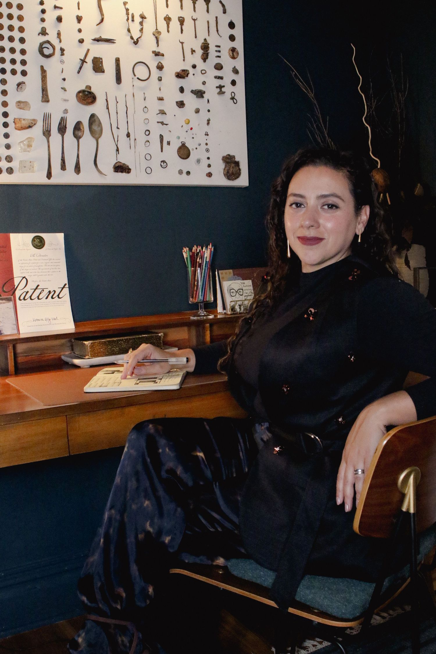 Woman in black satin dress sitting at vintage wooden desk in dark teal office space.