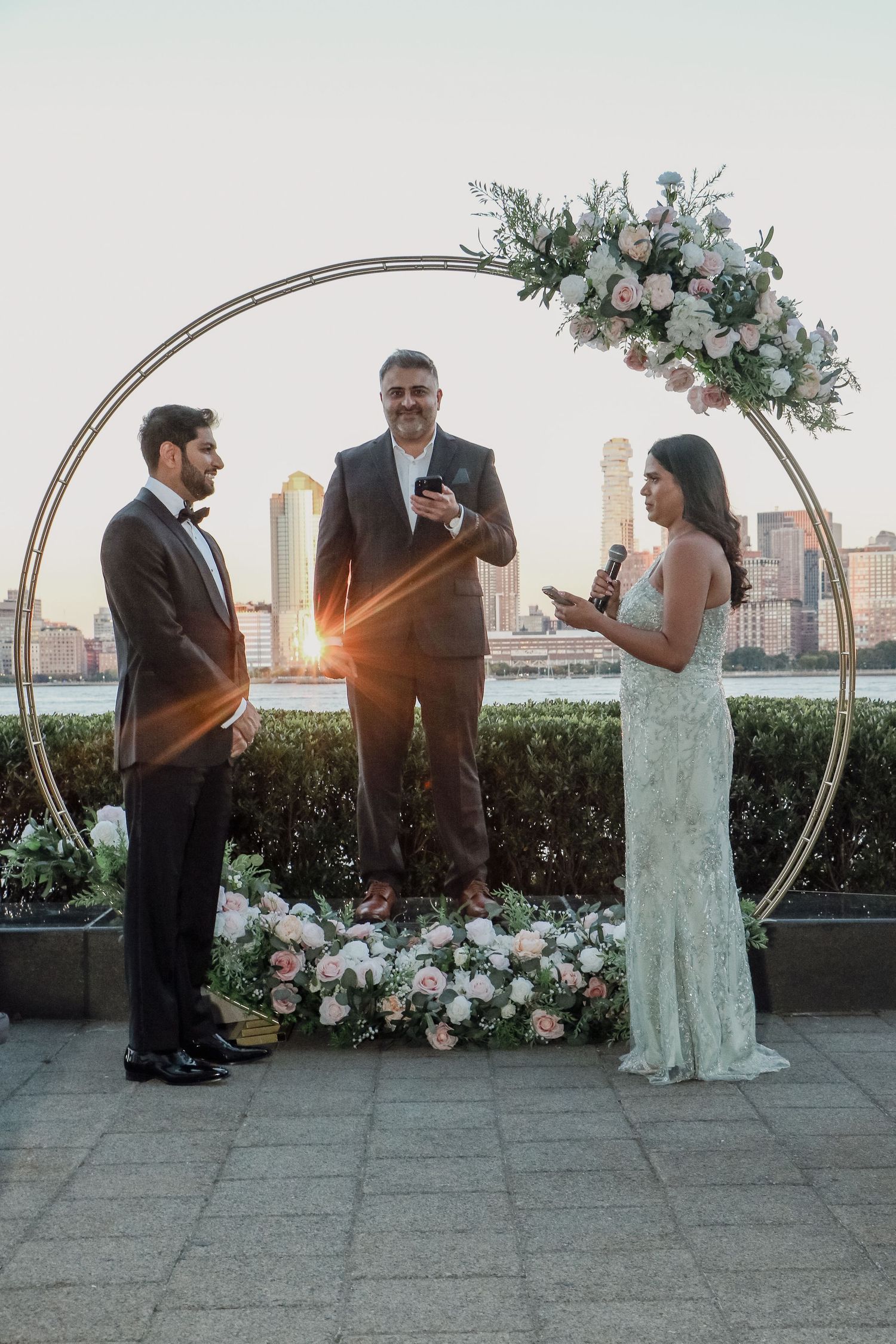 Couple exchanges vows at sunset under circular floral arch with NYC skyline in background.