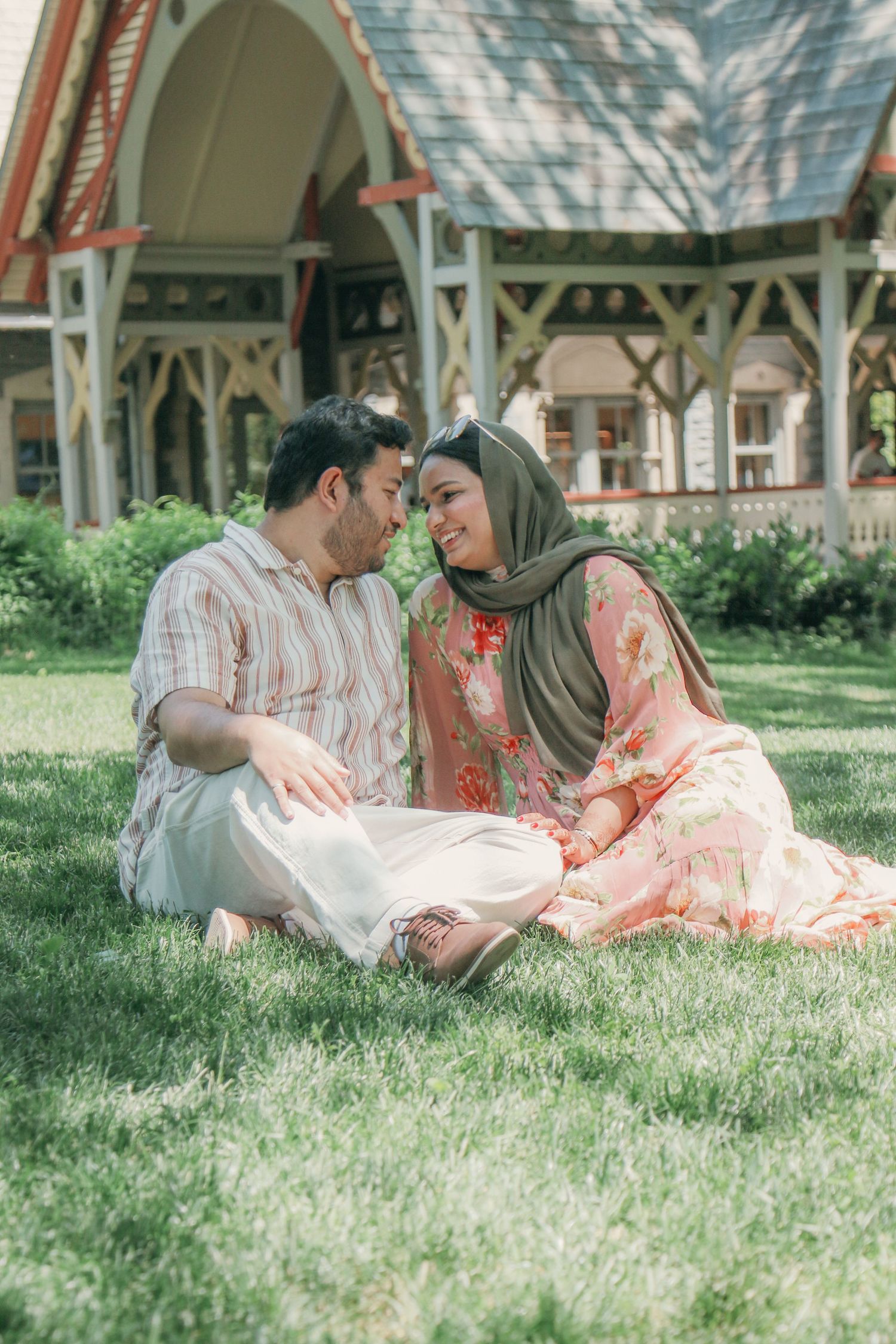 A couple in traditional attire sits on green grass, sharing an intimate moment in front of a charming wooden building.