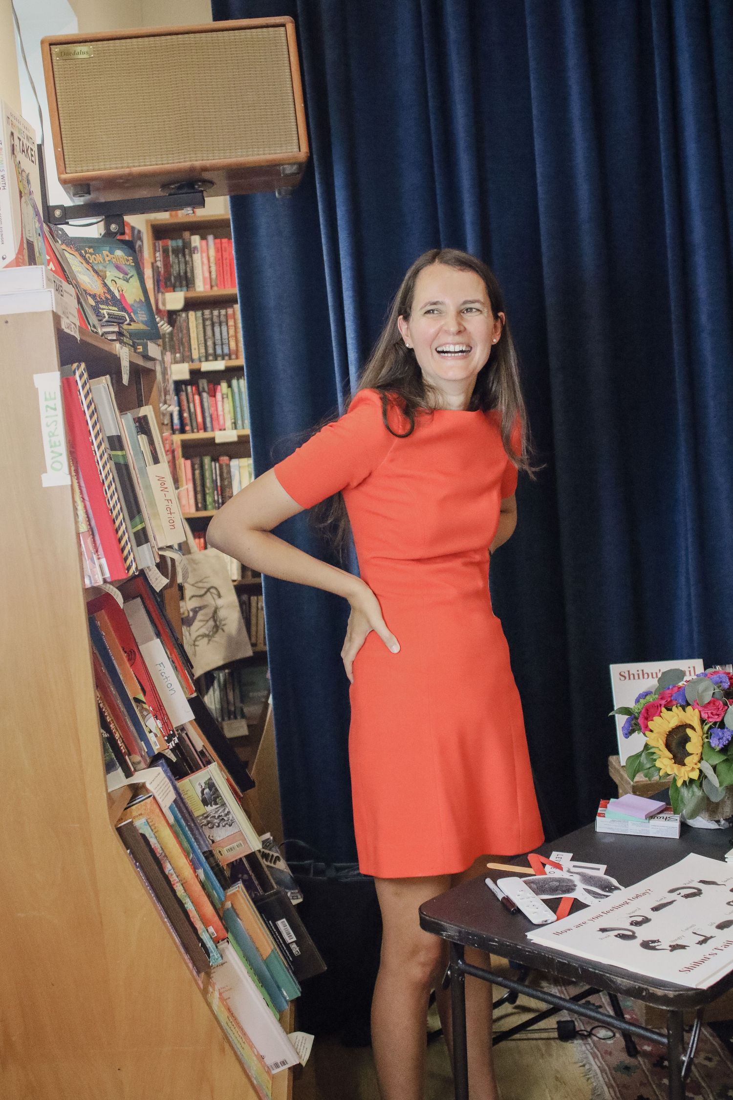 Woman in red dress laughing beside bookshelves at a book launch event.