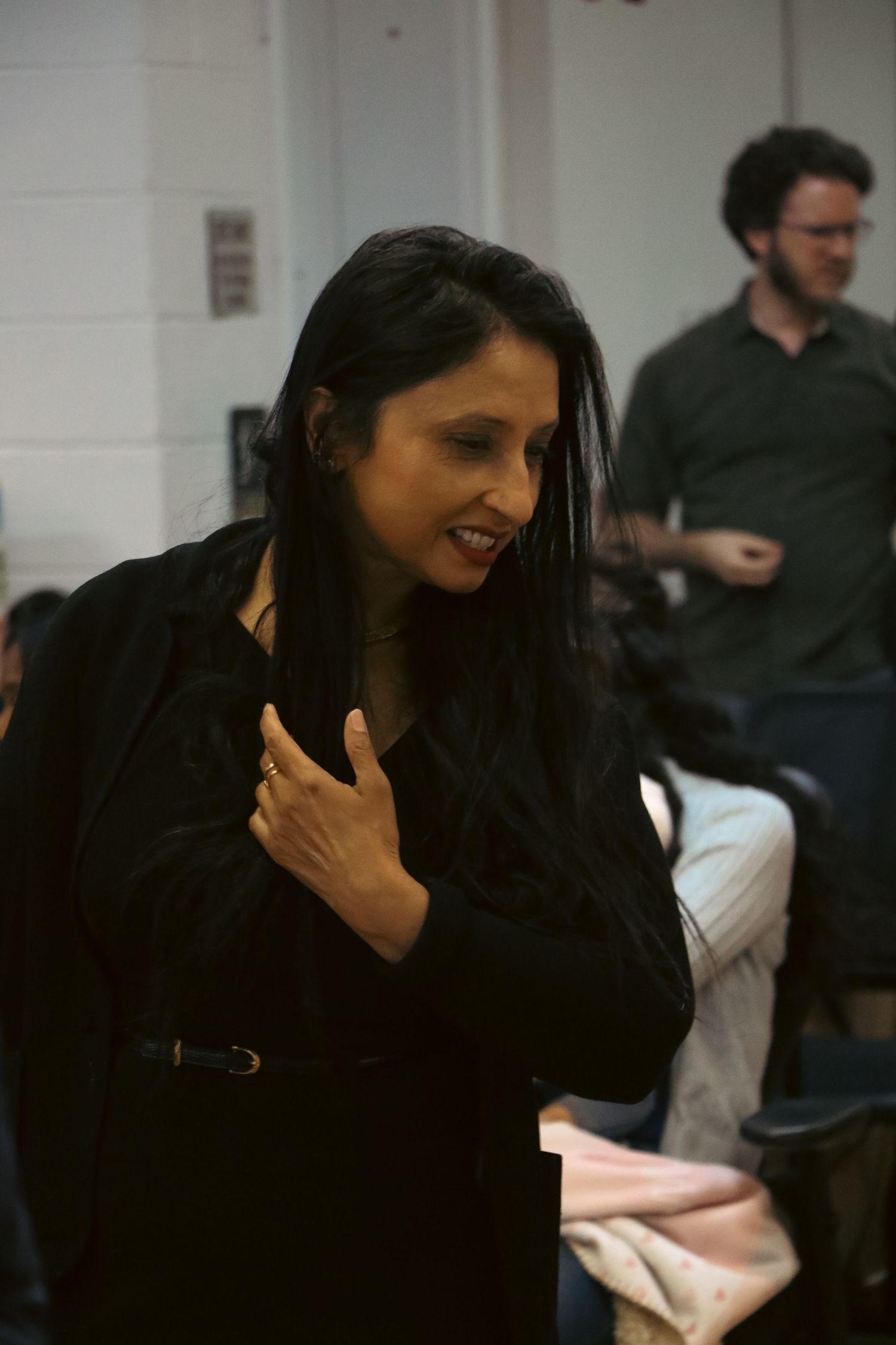 Woman in black jacket smiling at indoor event with people seated in background.