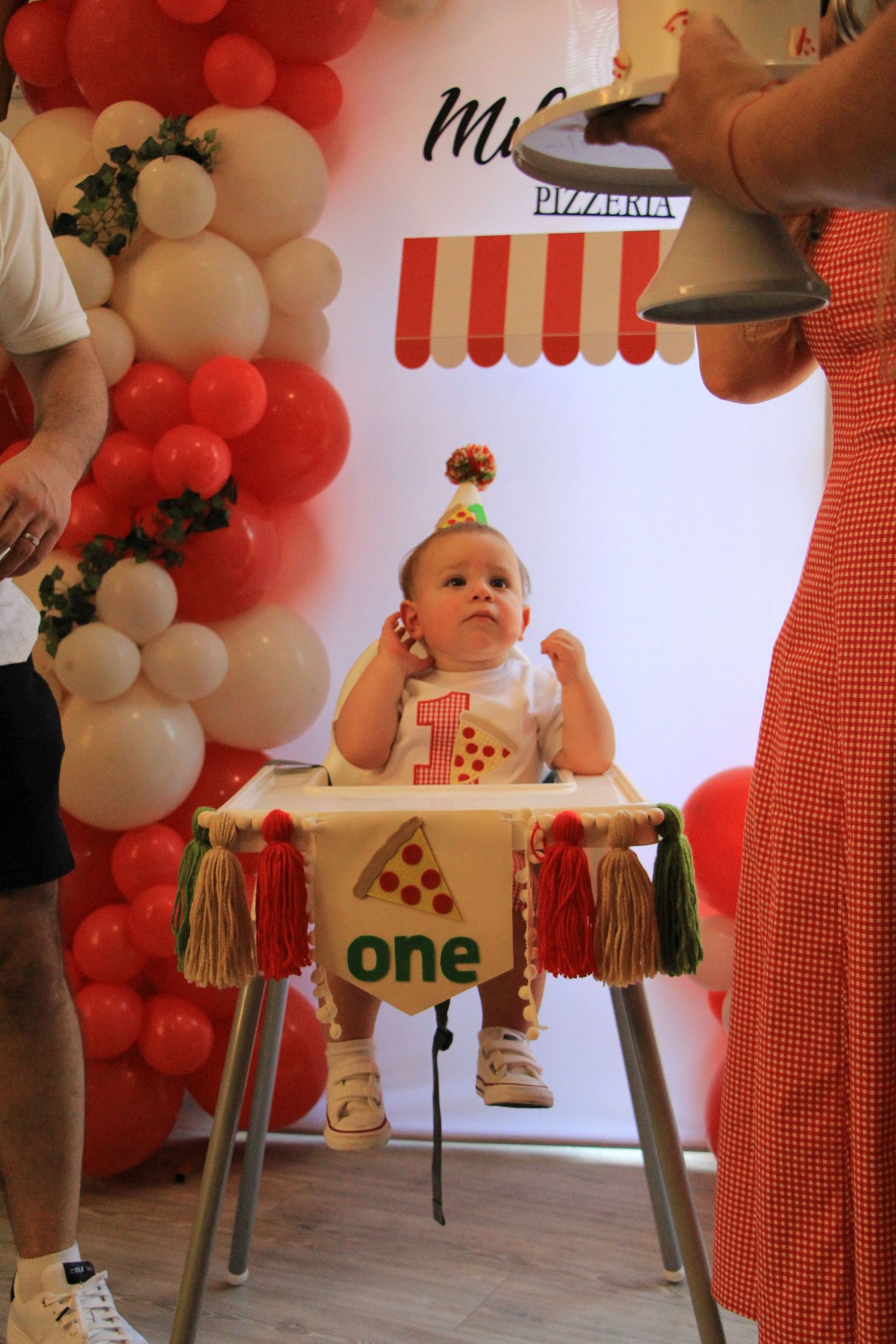 Baby in party hat sits in a pizza-themed high chair at Miles Pizzeria first birthday celebration.