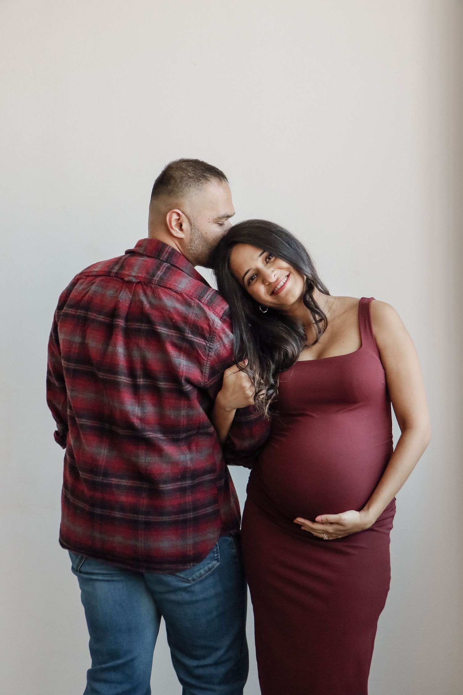 Pregnant woman in burgundy dress smiling as partner whispers in her ear.