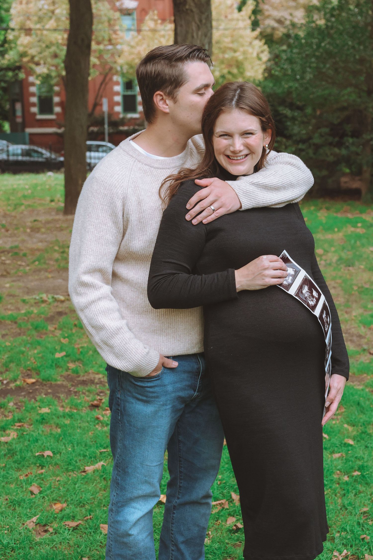 Smiling pregnant woman in a black dress holds ultrasound photos as her partner kisses her head in a sunny park with autumn leaves.