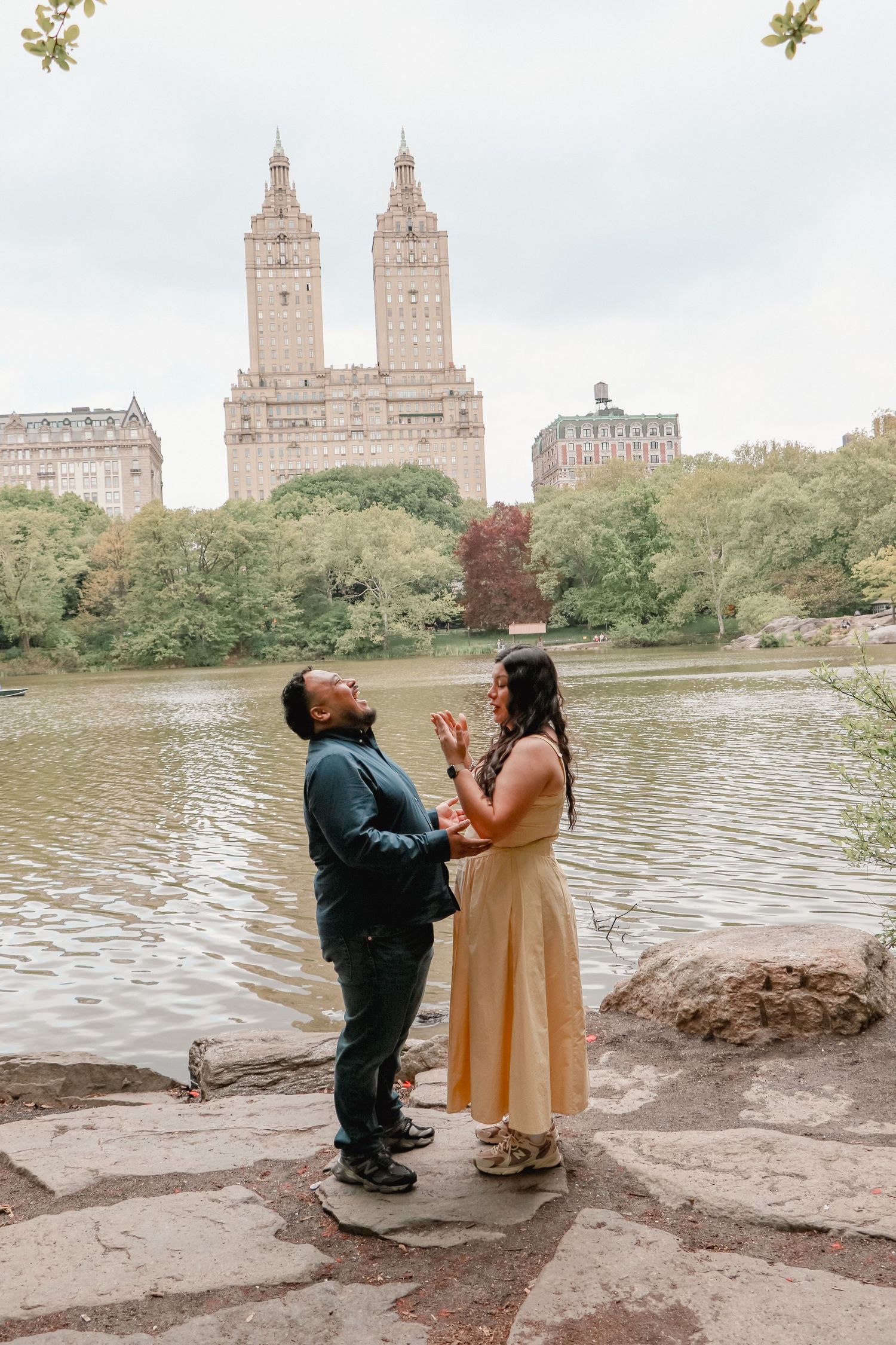 Couple sharing a joyful moment by the lake in Central Park, NYC, with the iconic San Remo building in the background.