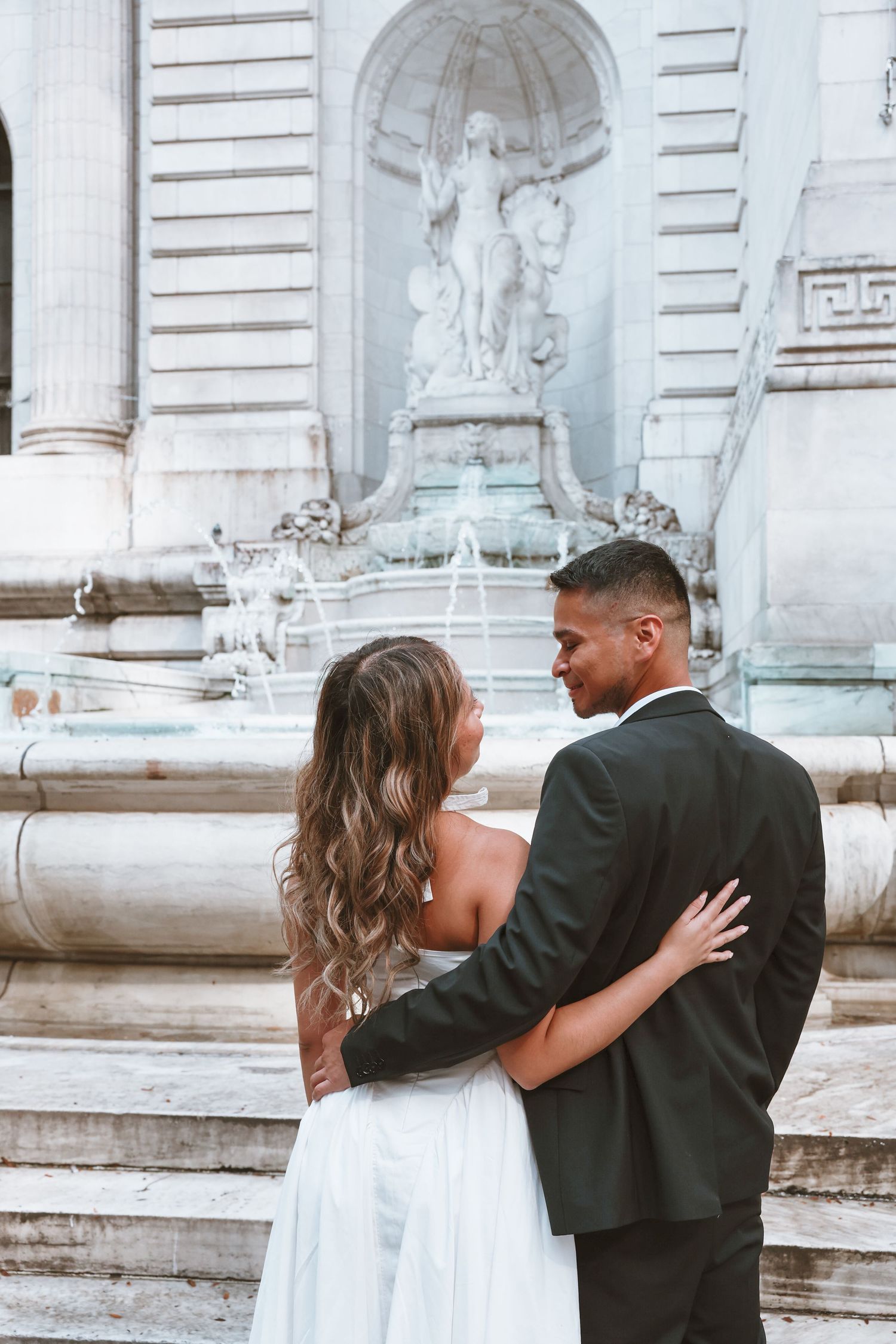 Couple embracing from behind in front of an ornate marble fountain with a statue, she in a white dress and he in a dark suit.