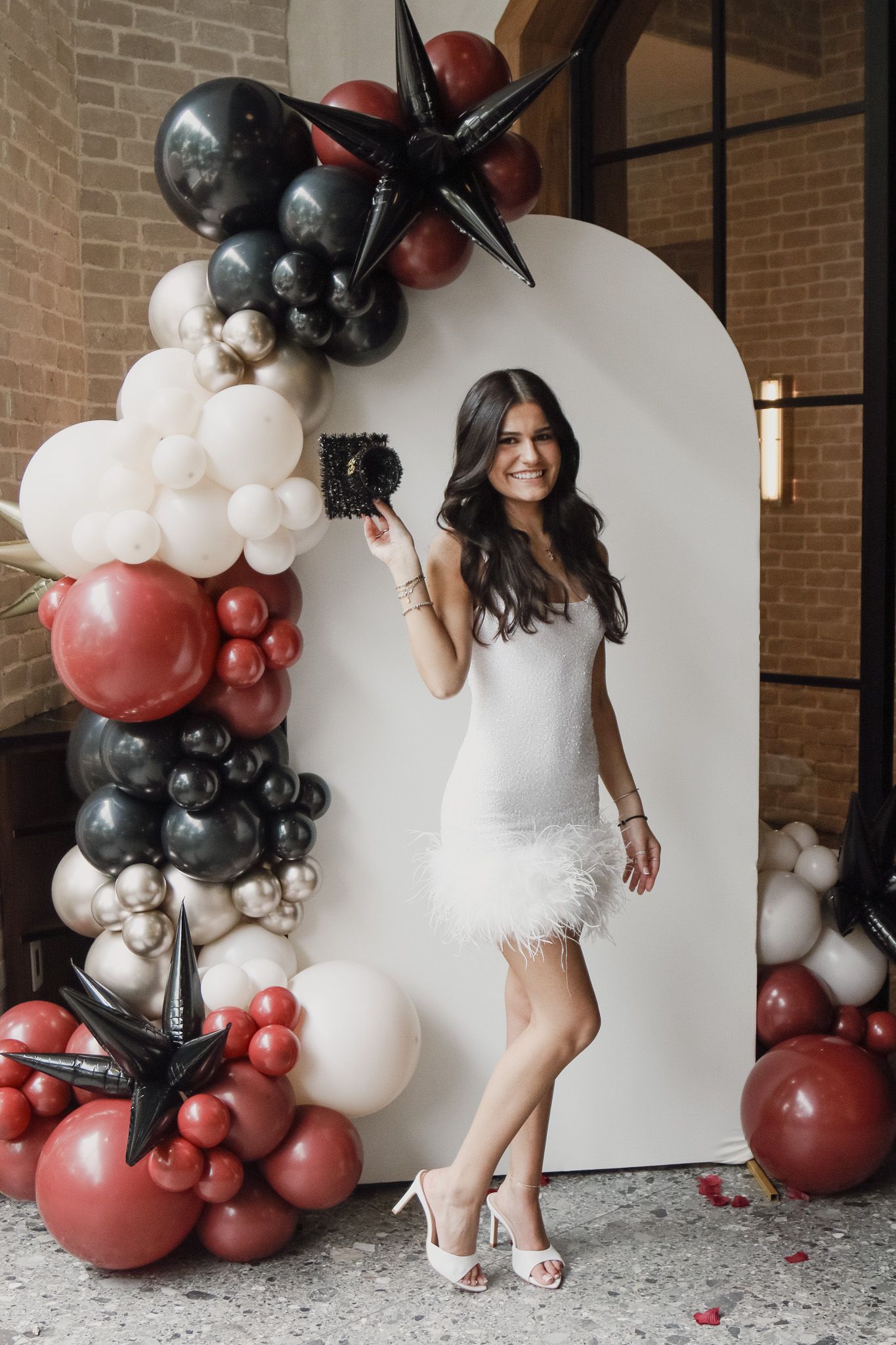 Smiling woman in a white feathered mini dress and heels poses with a camera in front of a black, red, and white balloon arch backdrop.