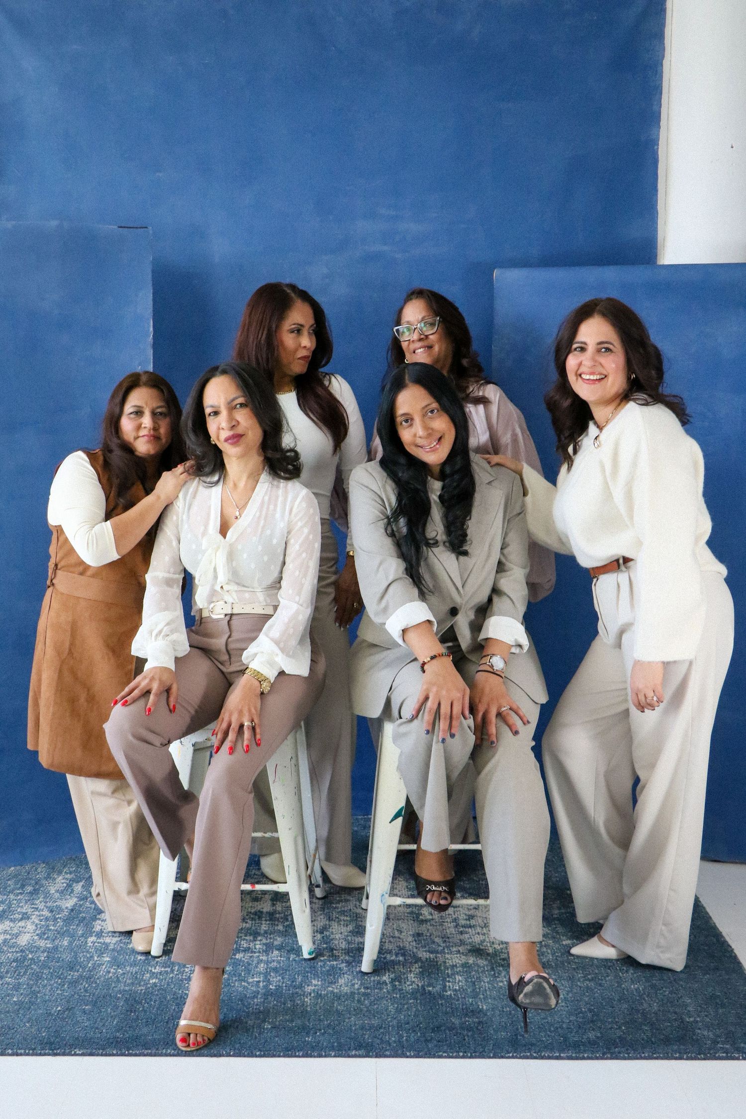 Group of six women posing together in neutral business attire against a blue painted studio backdrop.