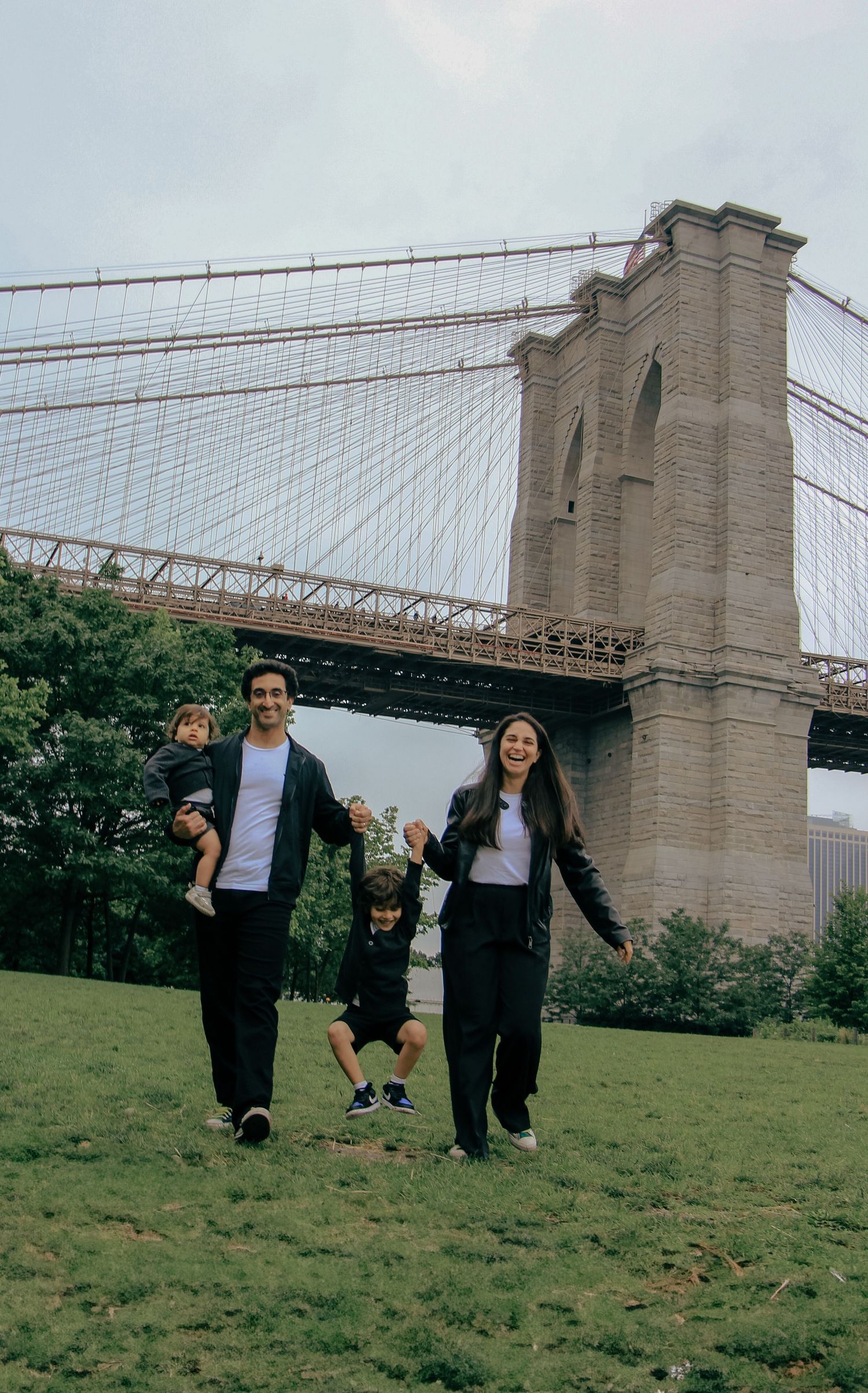 Family of four joyfully swinging child in front of Brooklyn Bridge on a green lawn.