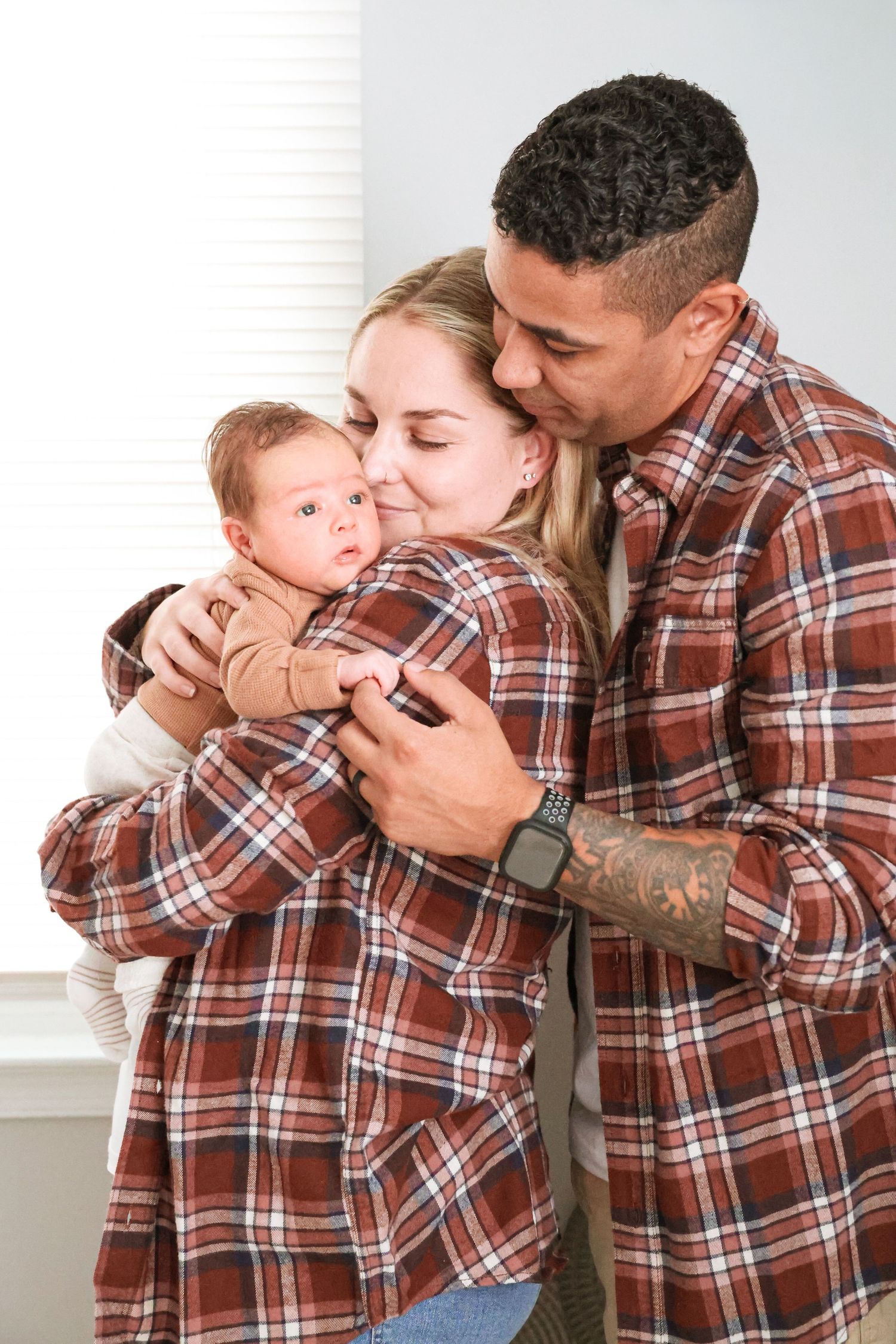 Smiling couple in matching red plaid flannel shirts embracing and cuddling their newborn baby in a bright, cozy room.