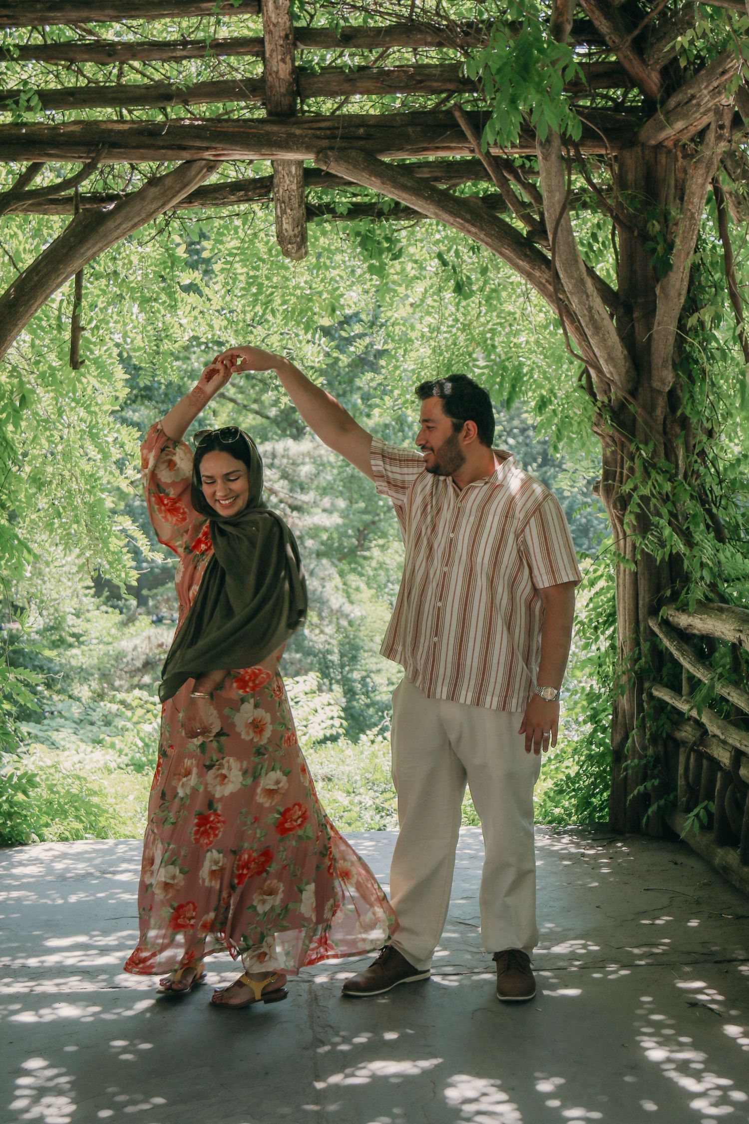 Couple dancing under a rustic wooden pergola surrounded by lush green trees in a garden setting.