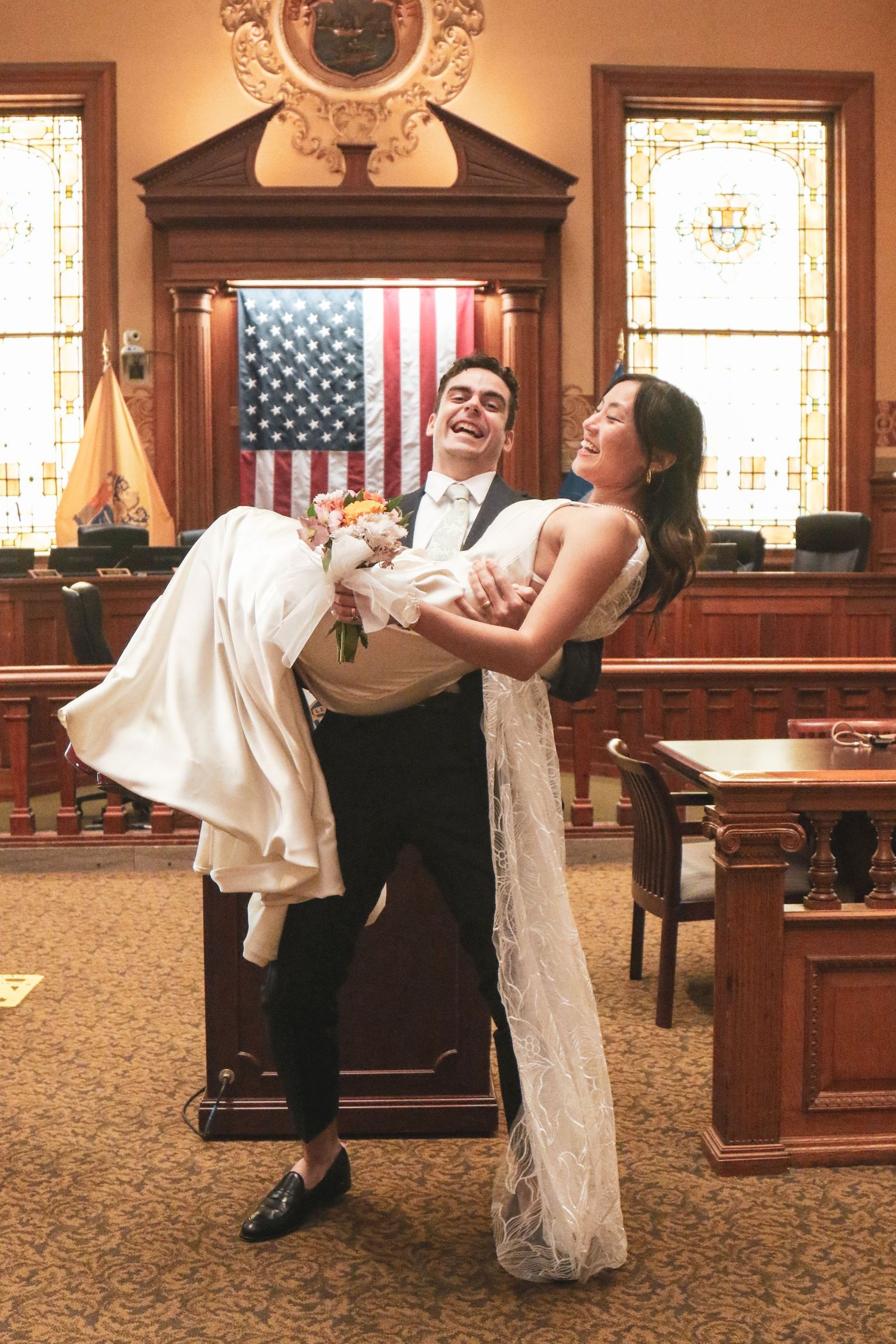 Groom lifting bride in wedding attire inside a courtroom with American flag backdrop.