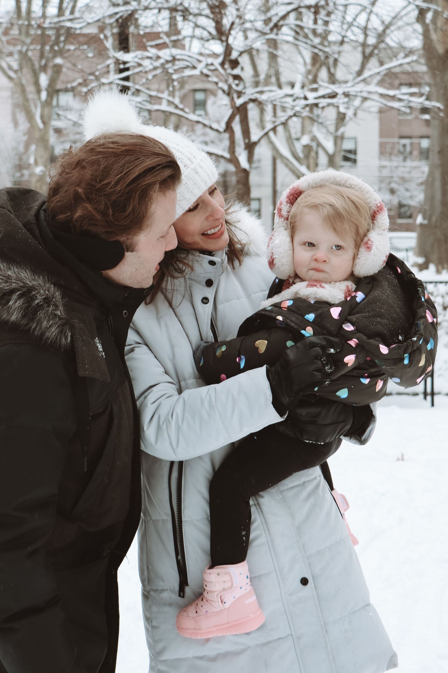 Parents holding their baby girl outdoors in a snowy park during winter.
