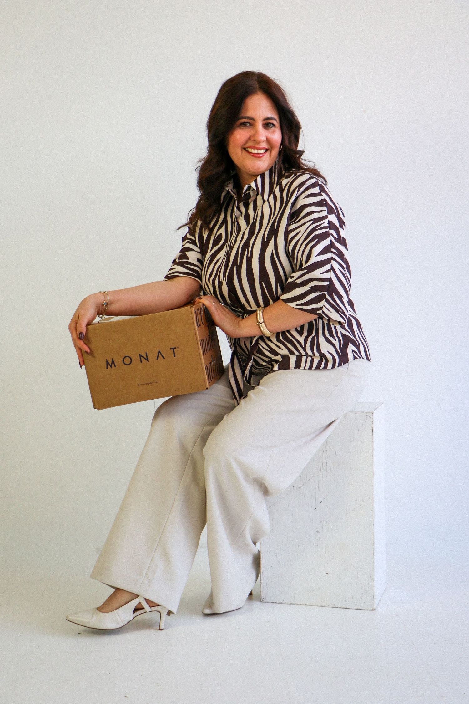 Smiling woman in zebra print shirt holding a MONAT box while seated on white block.