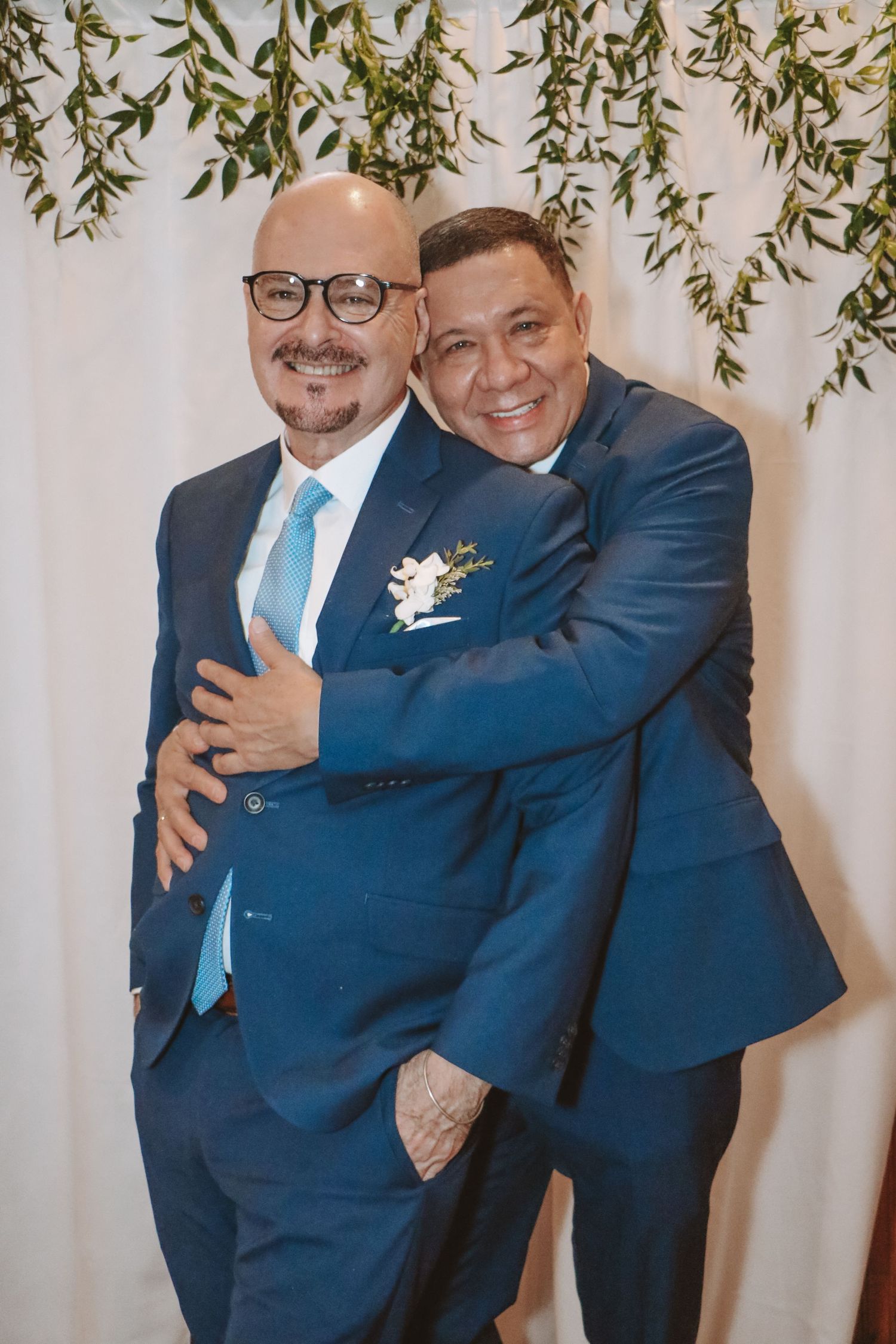 Two men in blue suits embracing warmly in front of a white floral backdrop with greenery at a wedding.