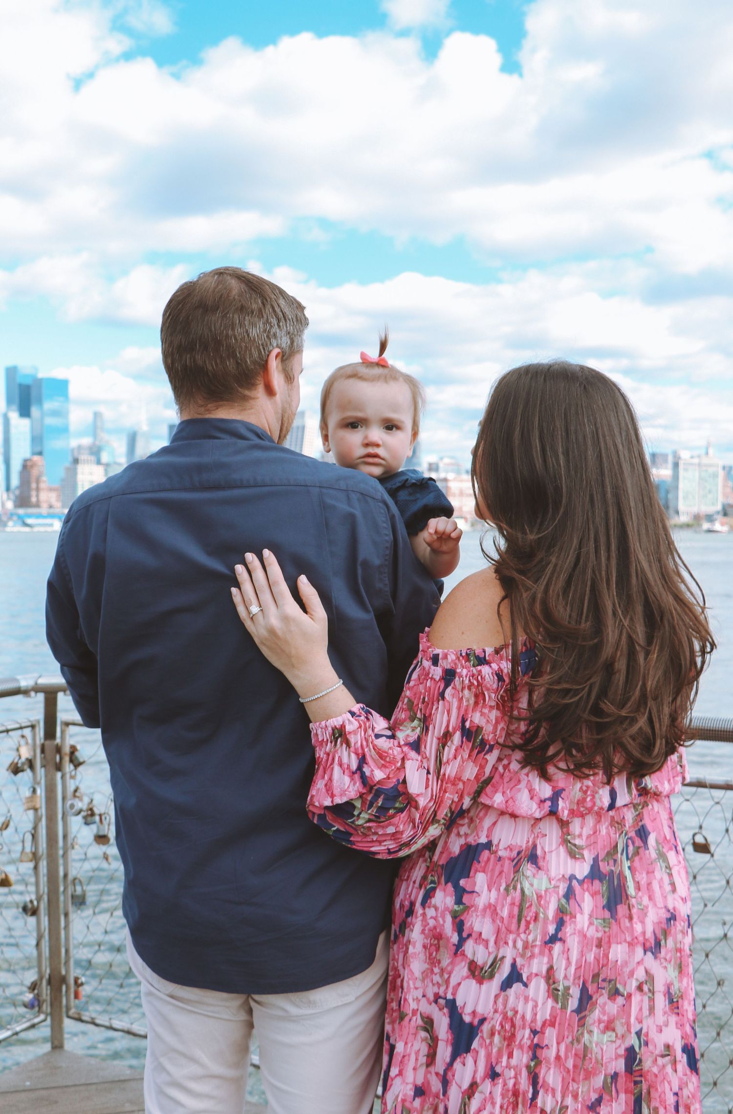 Parents holding baby facing New York City skyline from a waterfront dock with love locks.
