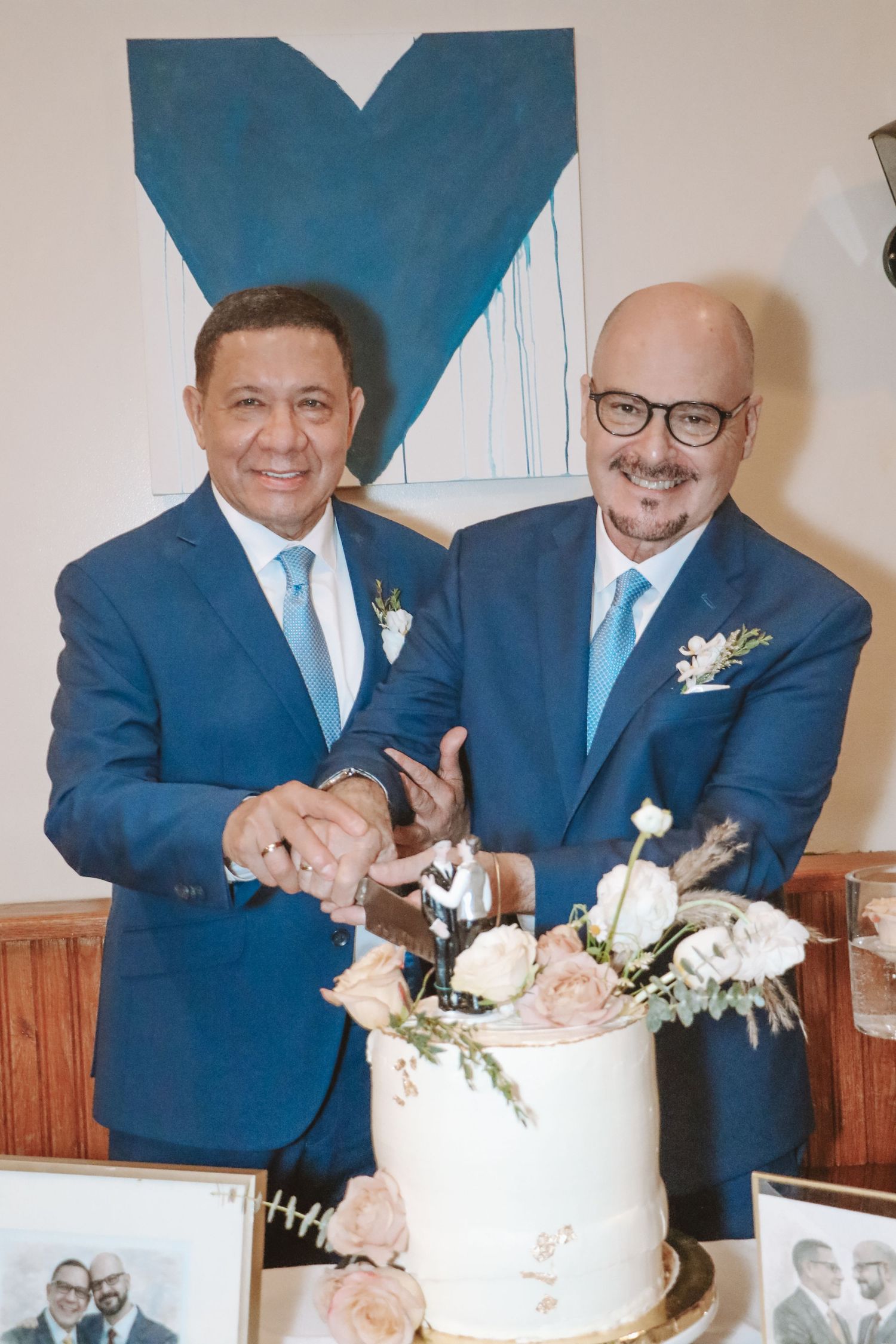 Two grooms in navy suits with blue ties cutting their floral wedding cake together.