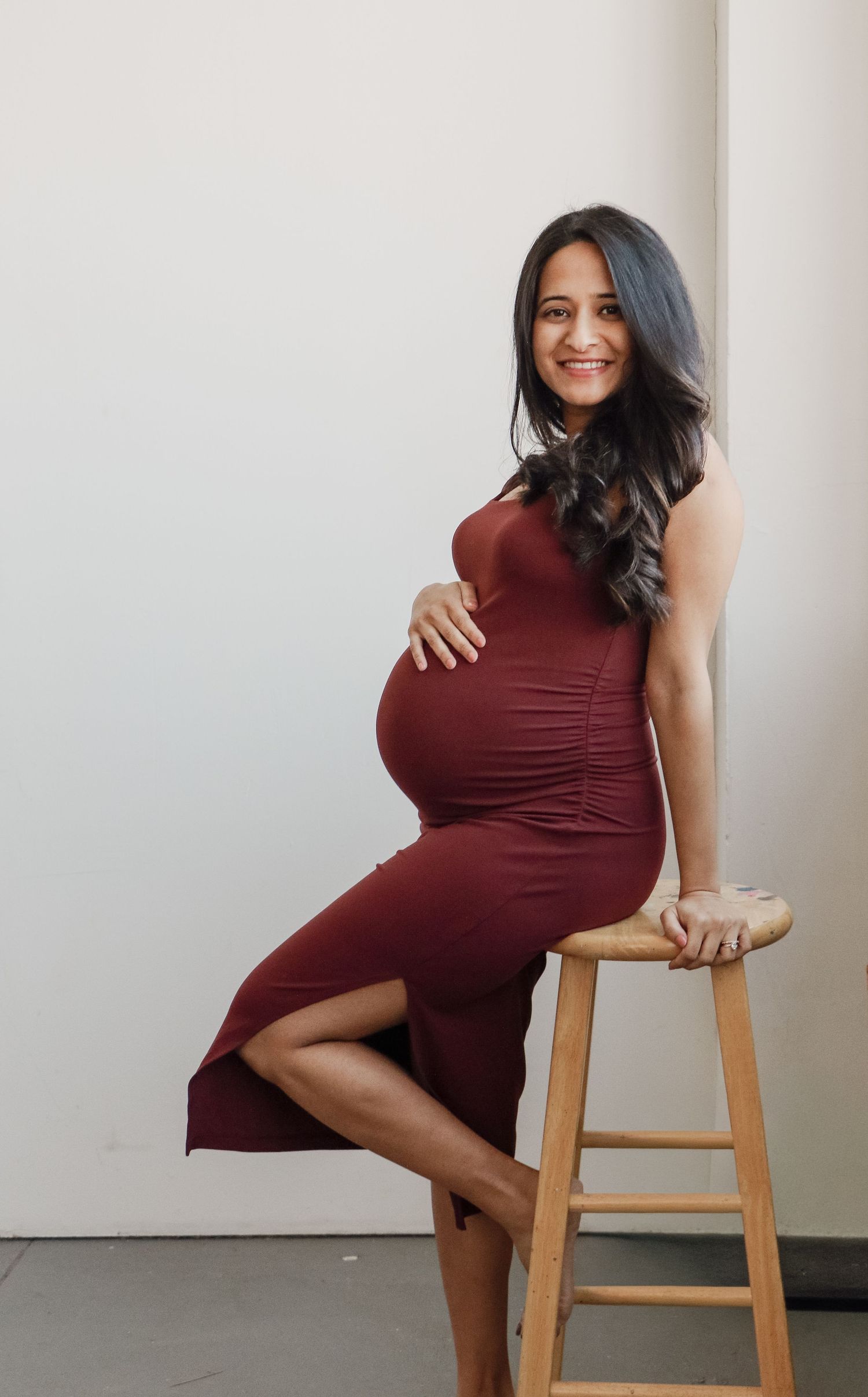 Smiling pregnant woman in burgundy dress poses on wooden stool in bright studio setting.