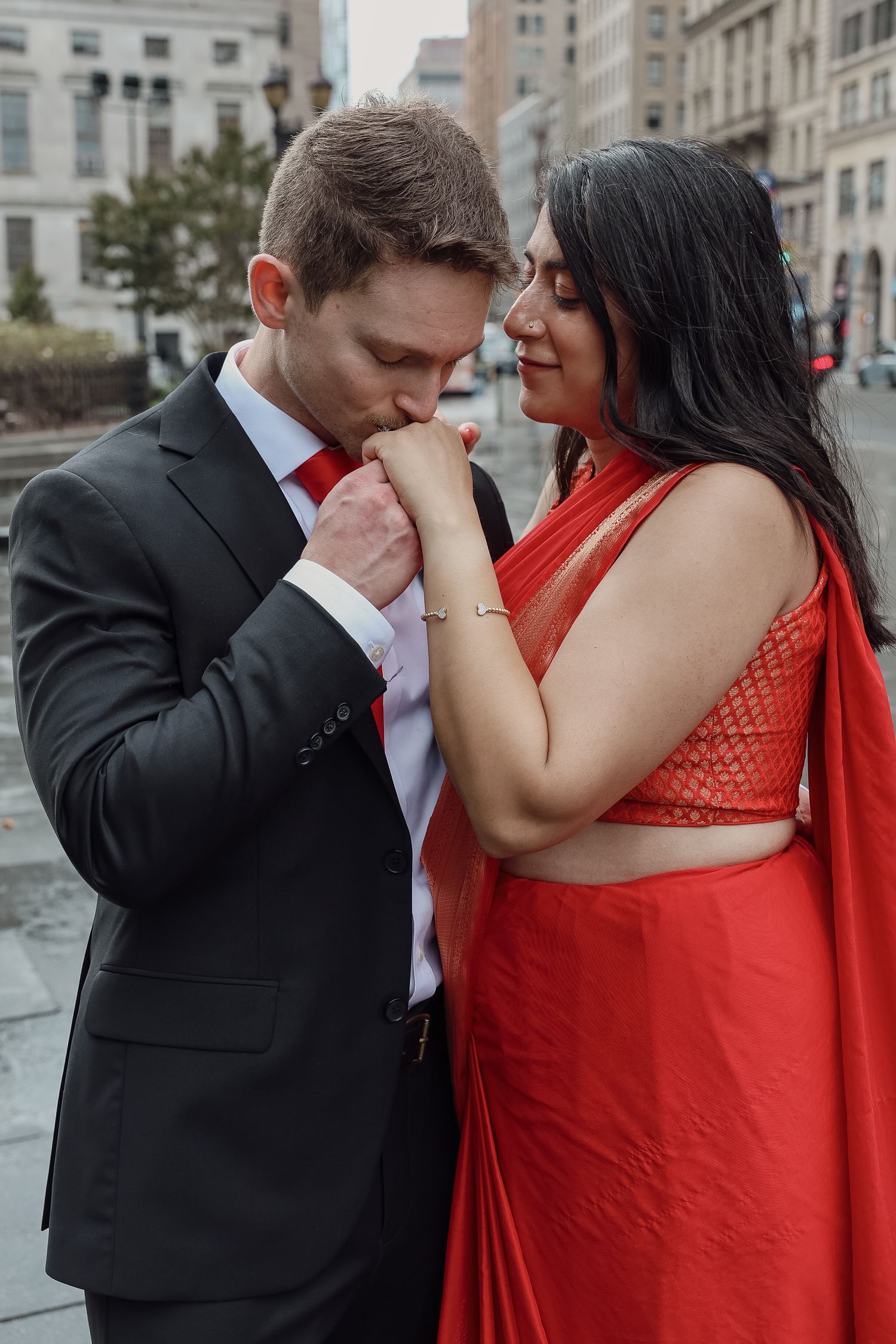 Man in a dark suit and red tie tenderly kisses the hand of a woman in an orange-red saree on a city street.